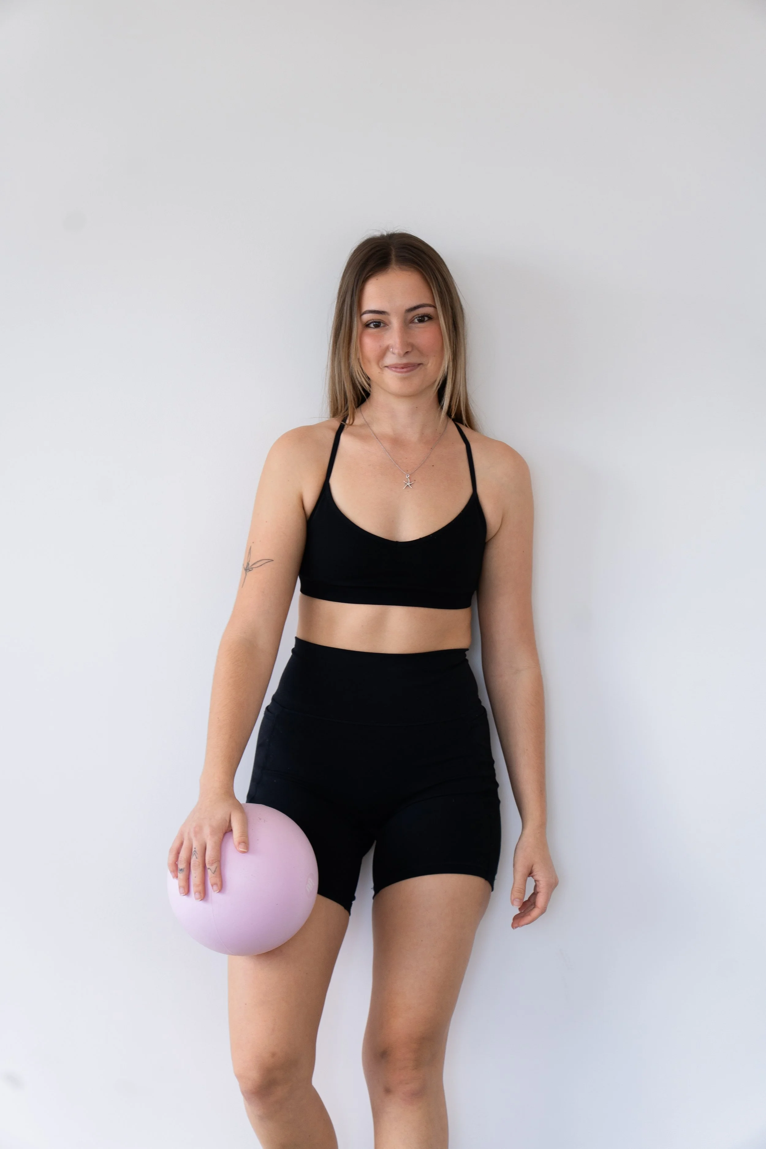 A young woman smiling, kneeling on an exercise mat while holding a green medicine ball, in a gym setting.