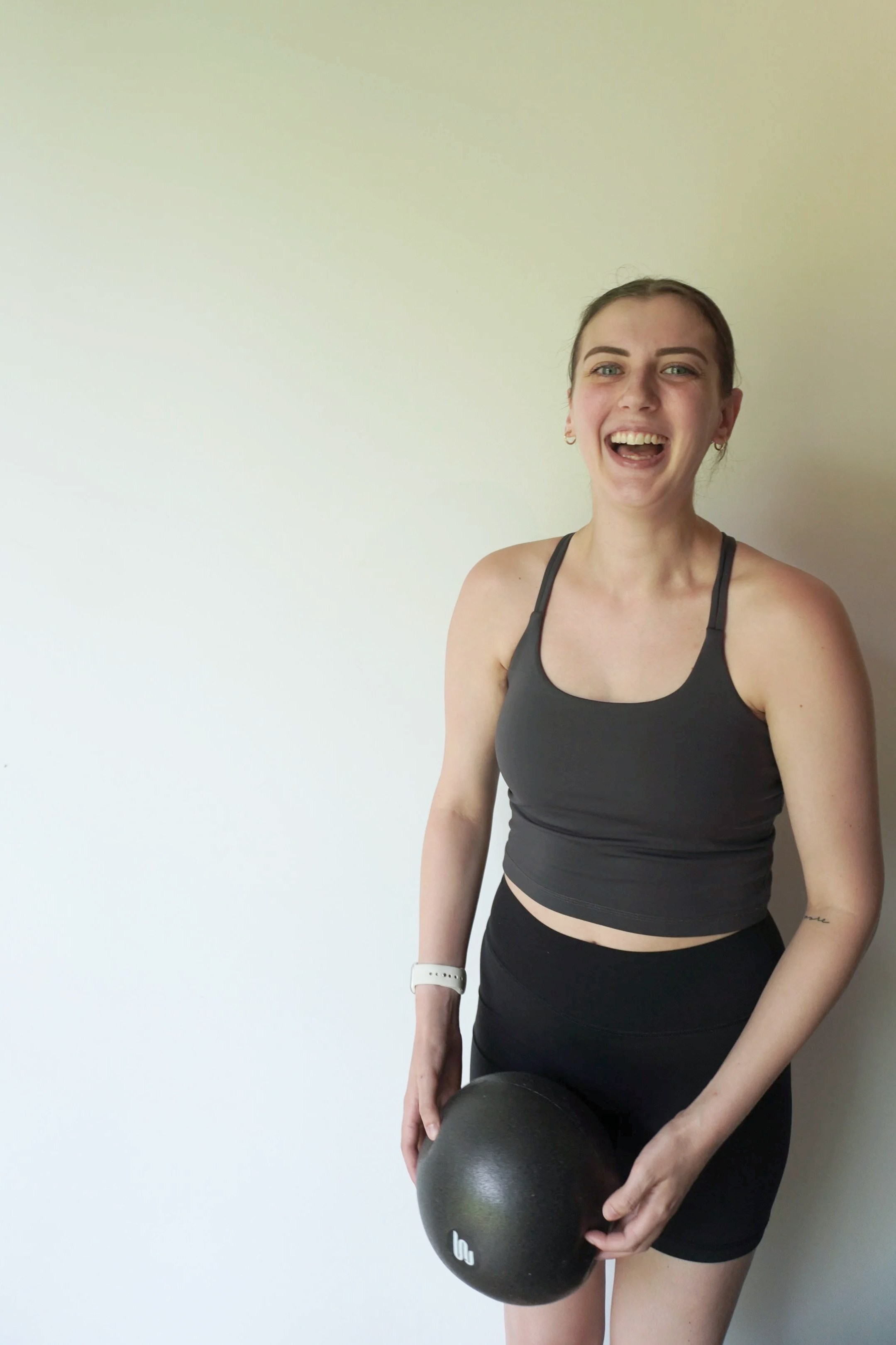 A young woman smiling and holding a black medicine ball, dressed in workout attire, standing against a blank wall.