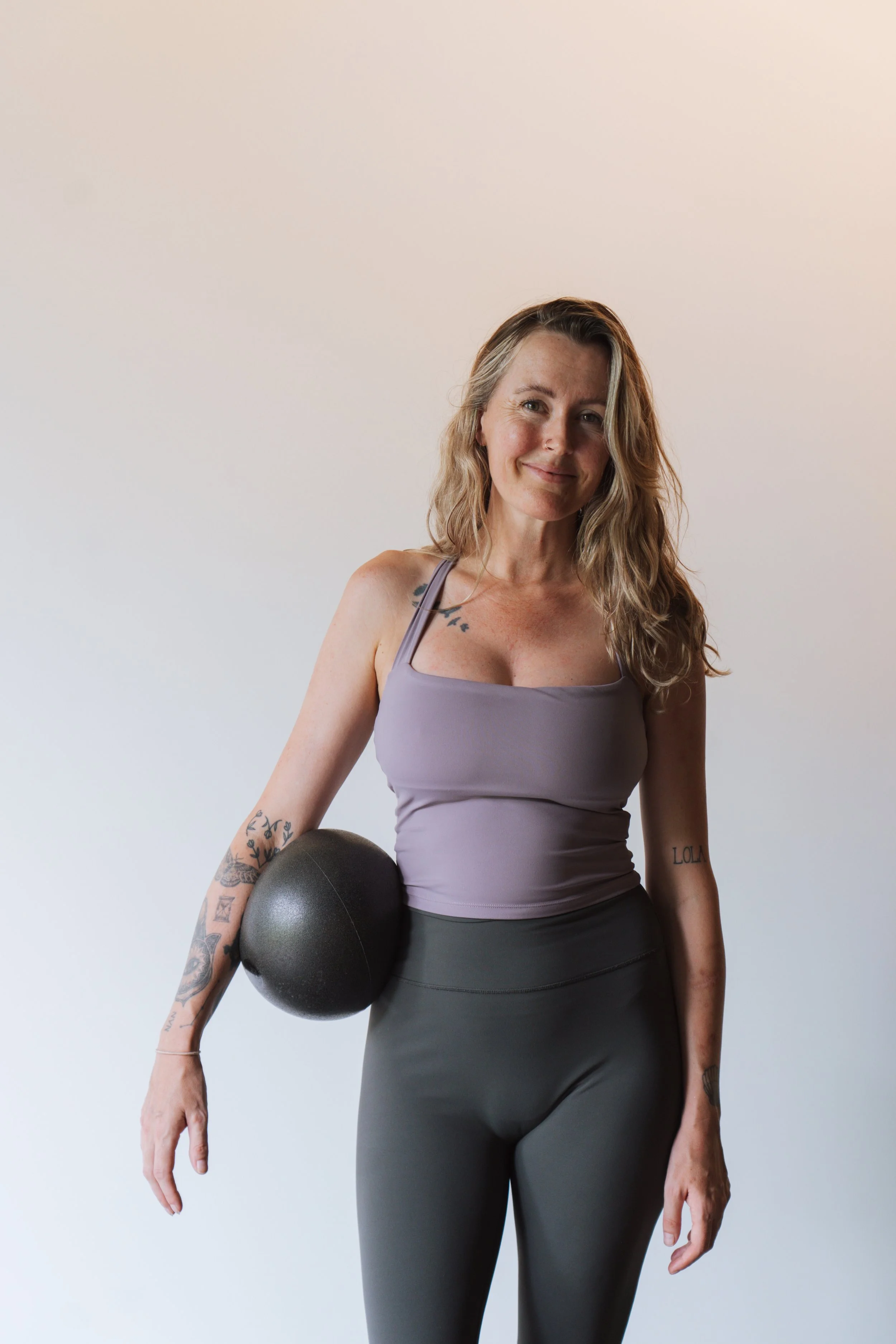 A pregnant woman with long wavy blonde hair and tattoos sits on a Pilates reformer machine, smiling gently, wearing a black tank top and black shorts, against a plain white background.