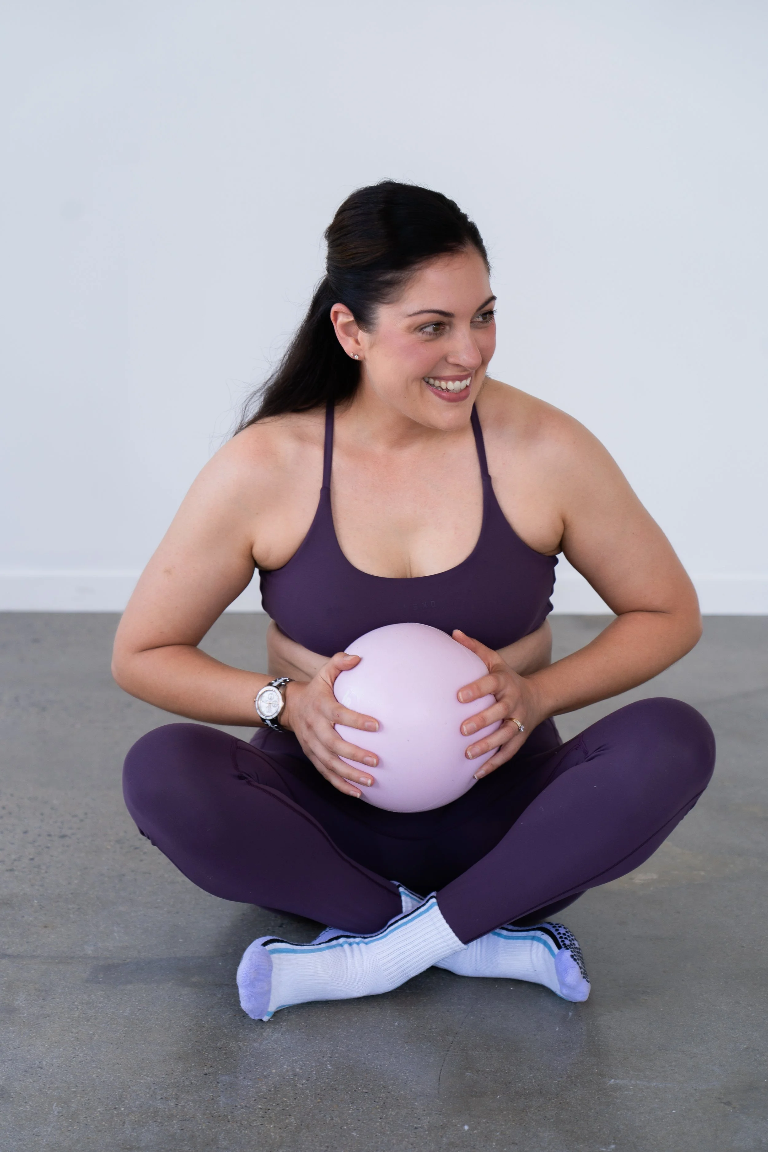 A woman sitting cross-legged on a Pilates reformer machine, smiling, with her arms raised, tossing a black exercise ball into the air.