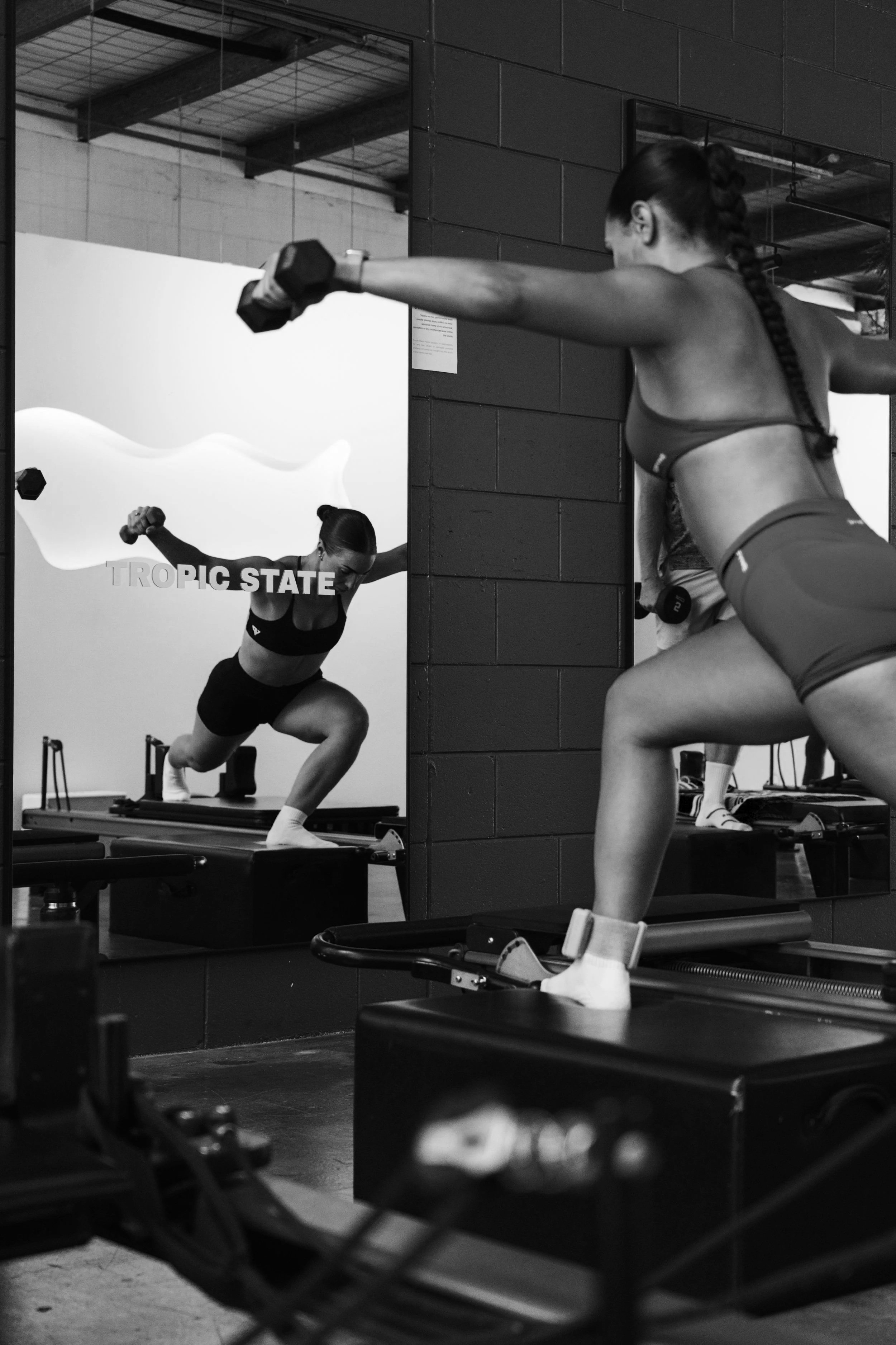 A woman in black athletic wear and white socks performs a squat with dumbbells in a fitness class, reflected in a mirror, with another woman in orange athletic wear participating, both using step platforms in a modern gym.