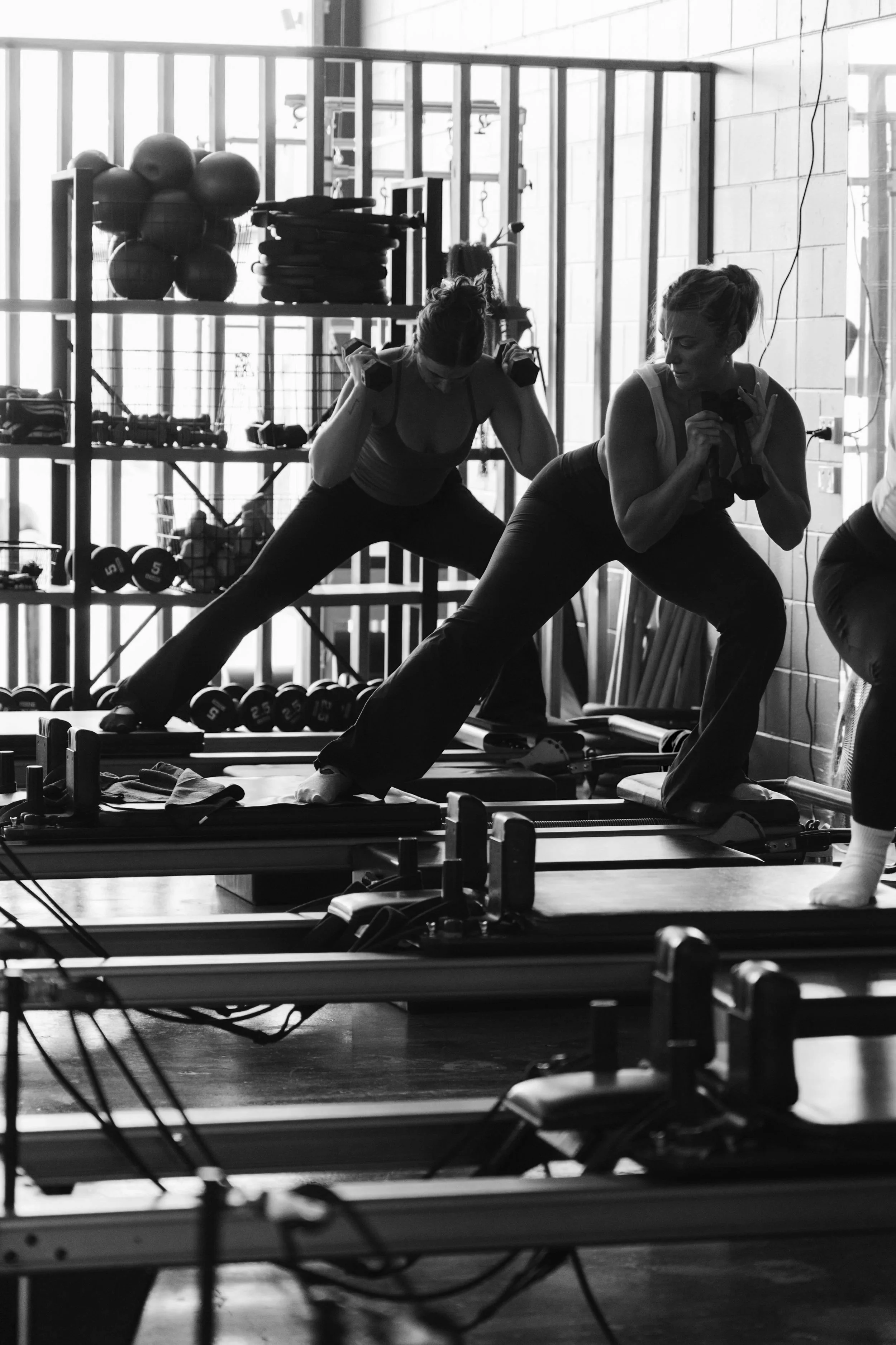 Two women exercising on reformer Pilates machines in a gym, holding dumbbells while performing strength exercise.