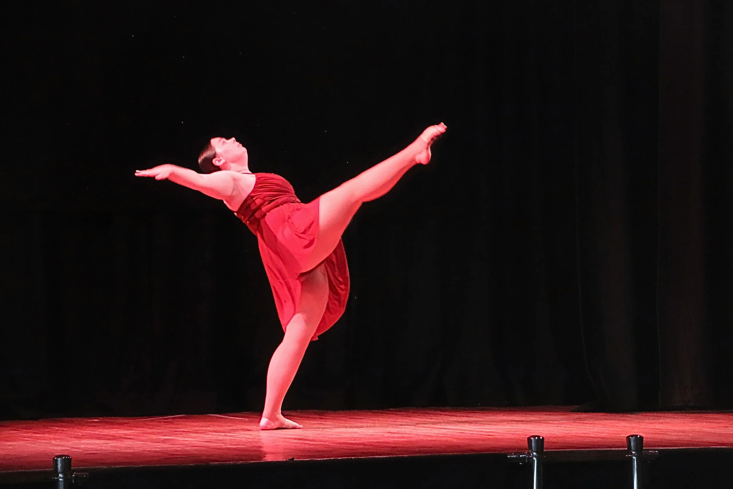 A Senior class dancer on stage in a red dress performing a jazz dance layout extension kick with one leg extended high and arms outstretched, against a black background during an LC Dance showcase performance.