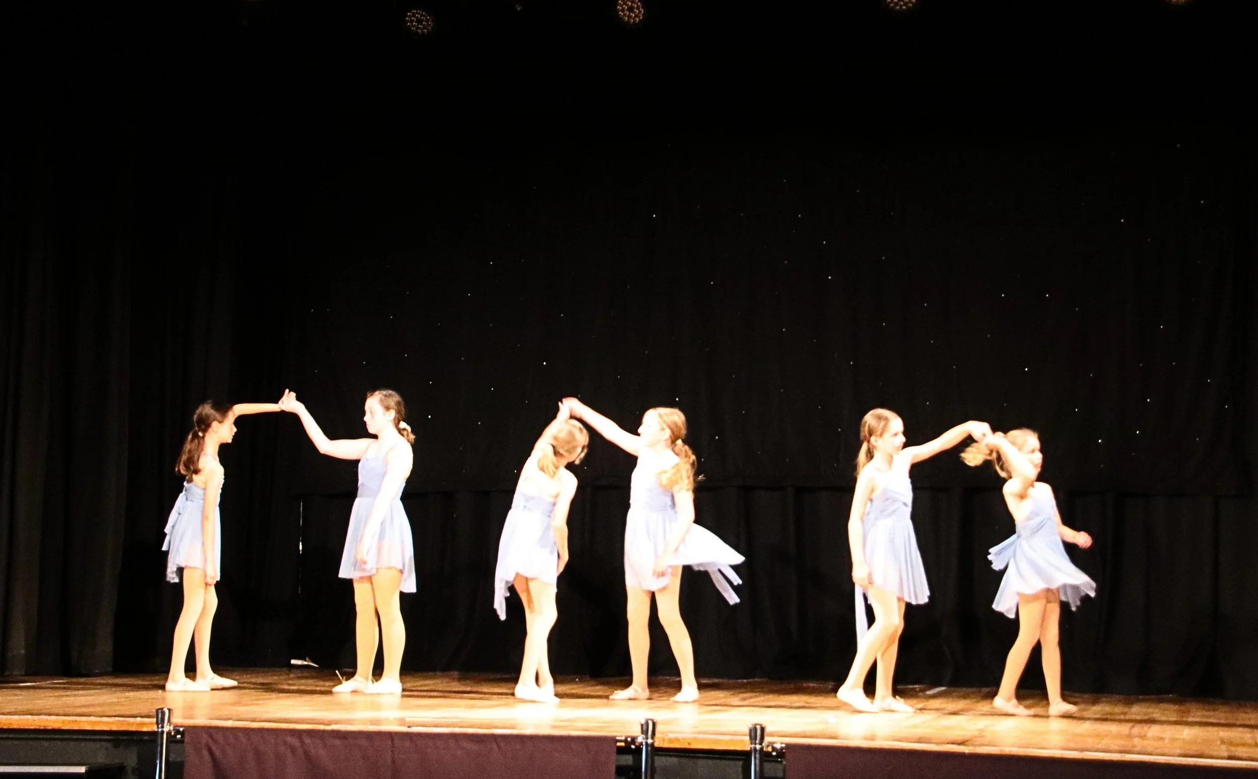 Six junior class students performing a lyrical dance on stage during an LC Dance showcase performance, holding hands and wearing light blue dresses, with a black backdrop and small star-like lights.