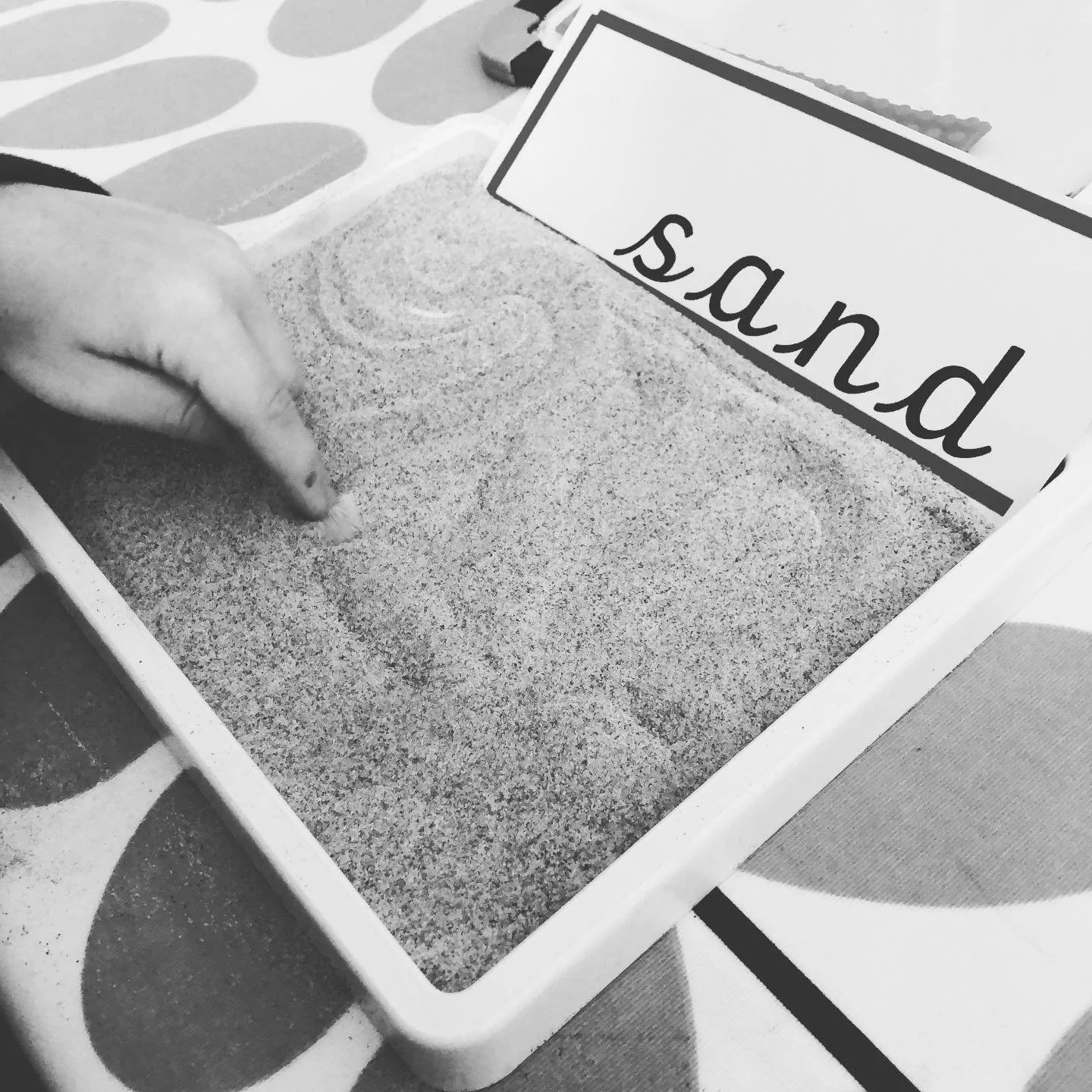 Person's hand sprinkling sand into a sand tray with a sign that reads 'sand' in the background, on a decorated table.