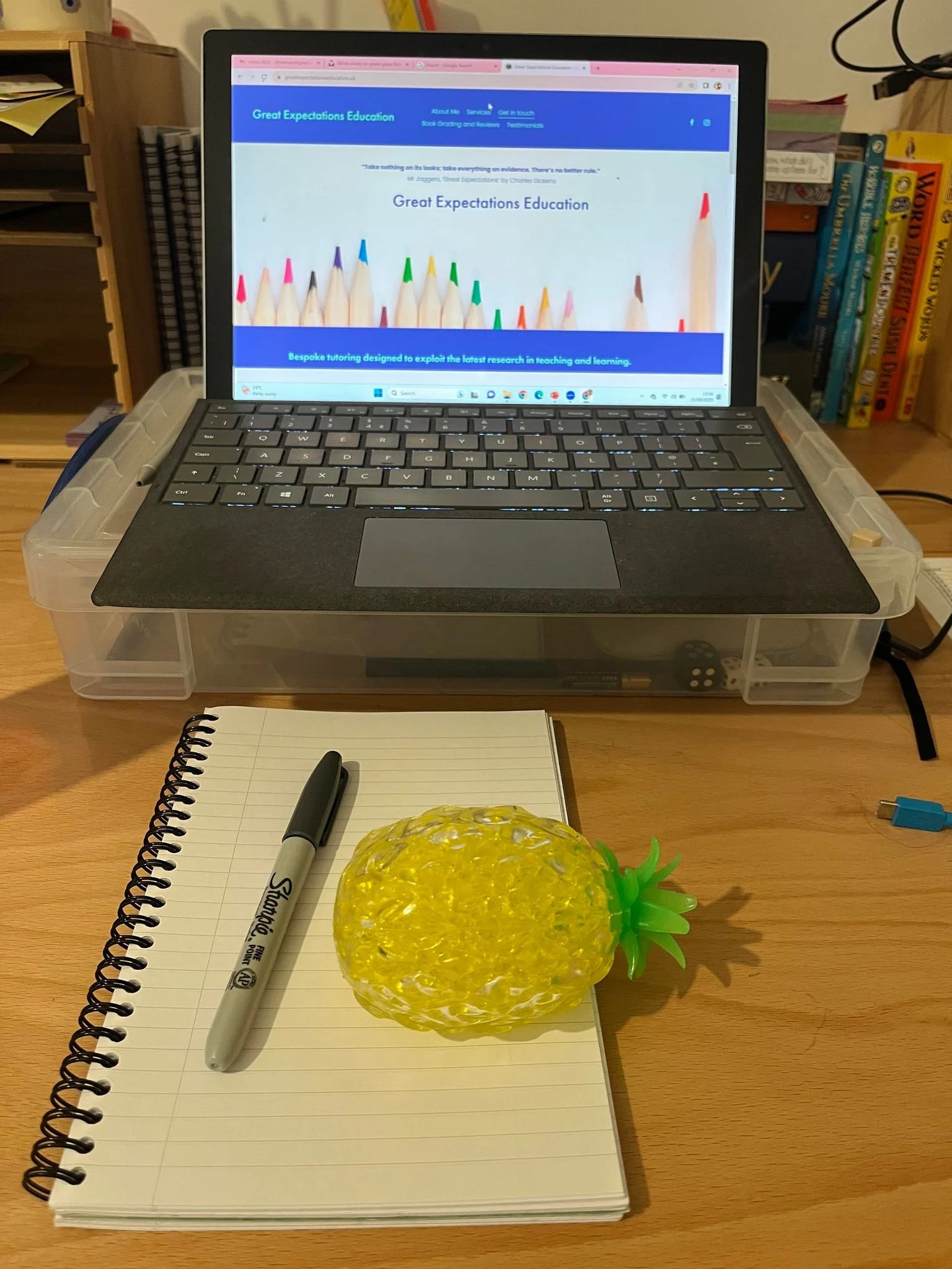 A desk setup with a transparent plastic container holding a laptop on top, a spiral notebook, a black Sharpie pen, and a yellow pineapple-shaped object on the notebook. There are books on shelves in the background and a blue cable on the right side.
