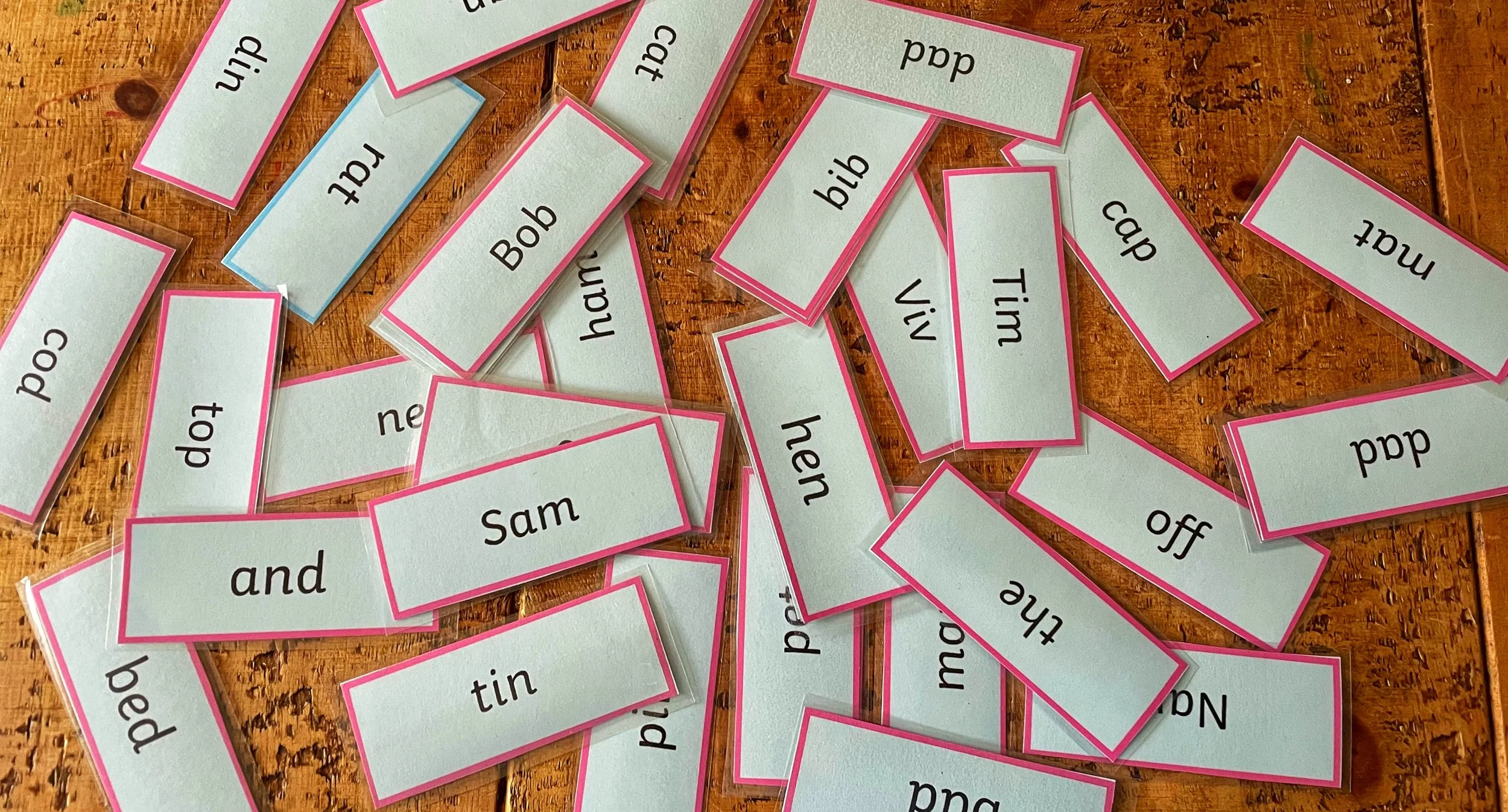 Spelling flashcards with words like 'and,' 'top,' 'off,' 'bad,' 'bed,' 'hat,' 'him,' 'tin,' 'look,' 'bib,' 'Vivi,' and 'Sam,' scattered on a wooden surface.