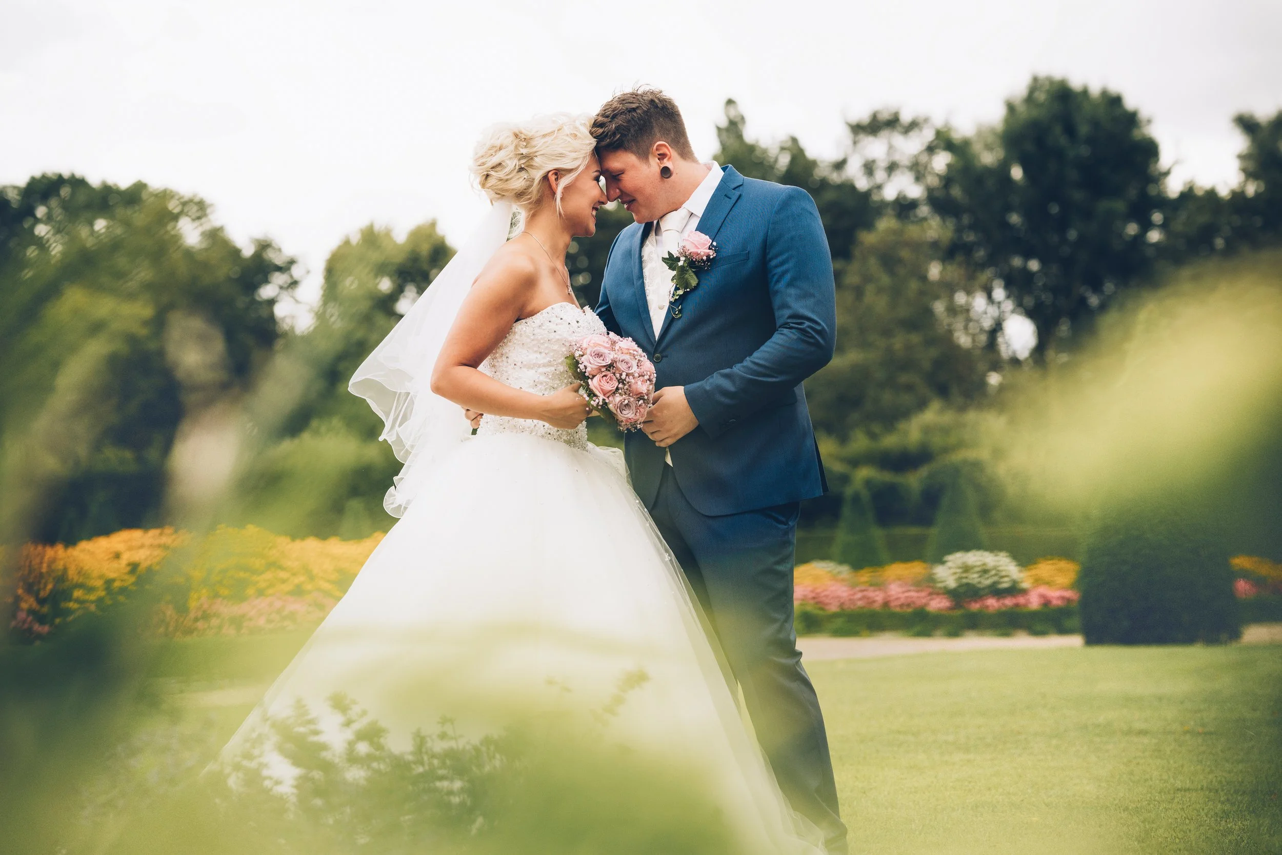 Ein Brautpaar steht im Freien, die Braut trägt ein weißes Hochzeitskleid und hält einen Blumenstrauß, der Bräutigam trägt einen blauen Anzug. Sie berühren ihre Stirn zärtlich und lächeln. Im Hintergrund sind Bäume und Blumen sichtbar.