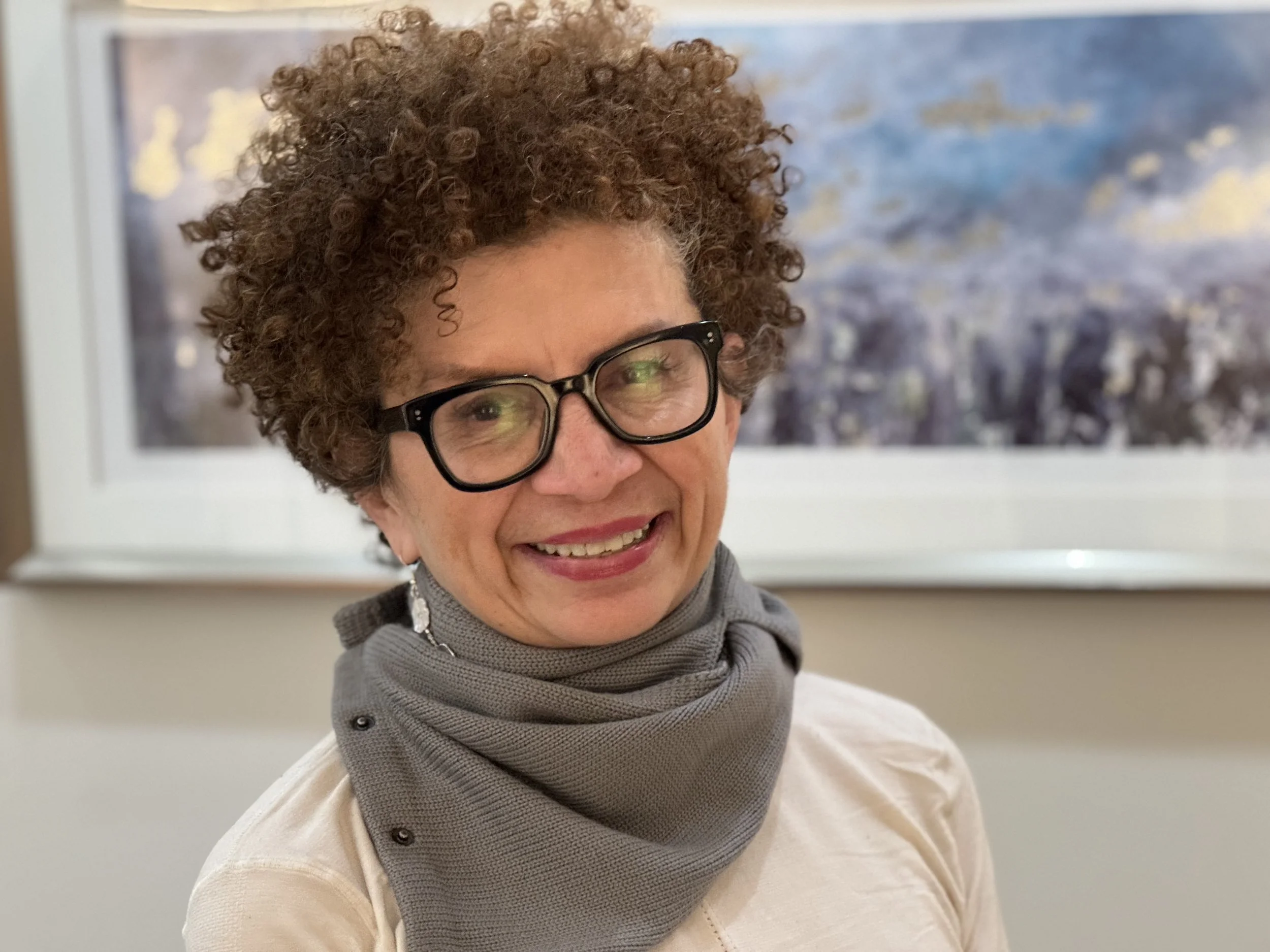 A woman with short curly hair, wearing glasses, smiling, and dressed in a light-colored shirt and gray scarf, standing in front of a window with a mountainous landscape painting in the background.