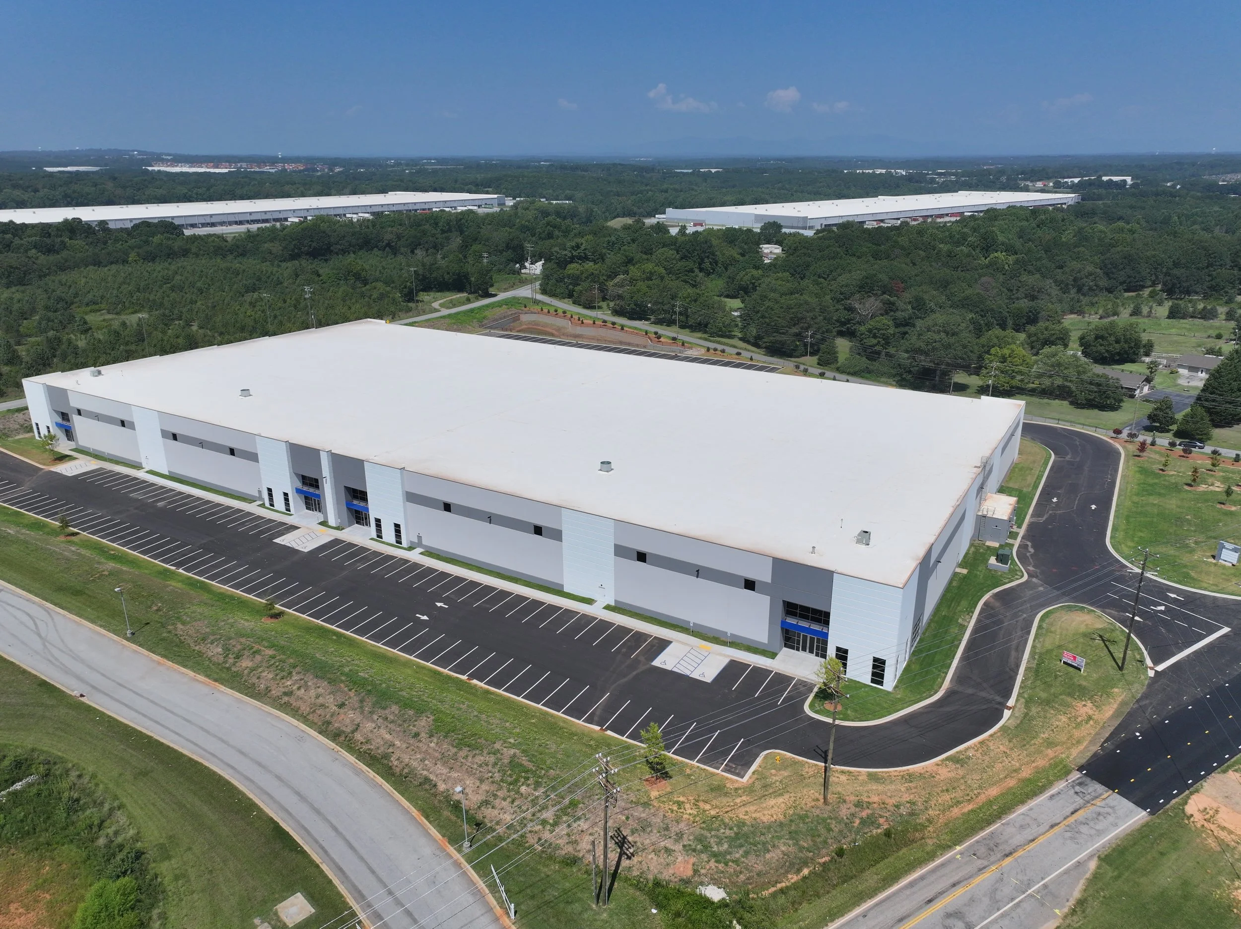 Aerial view of a large industrial warehouse building with a white roof and surrounding parking lot, set in a green landscape with trees and distant warehouses.