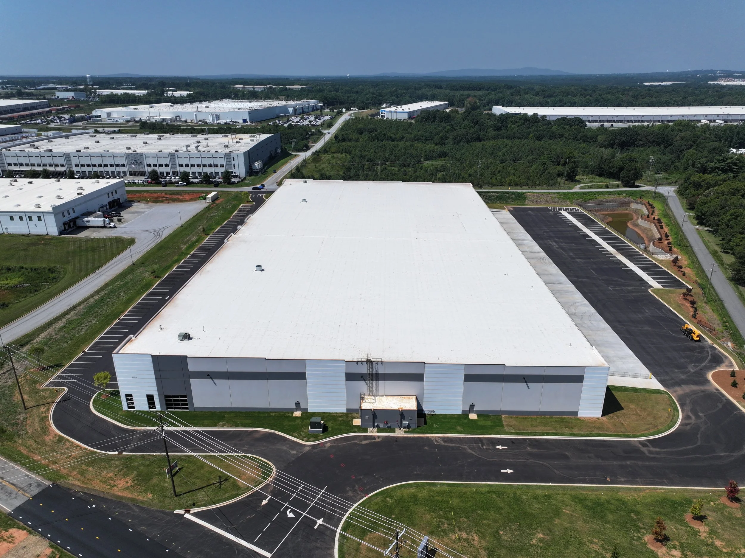 Aerial view of a large industrial warehouse with a white roof, surrounded by paved parking lots and greenery, located in an industrial park.