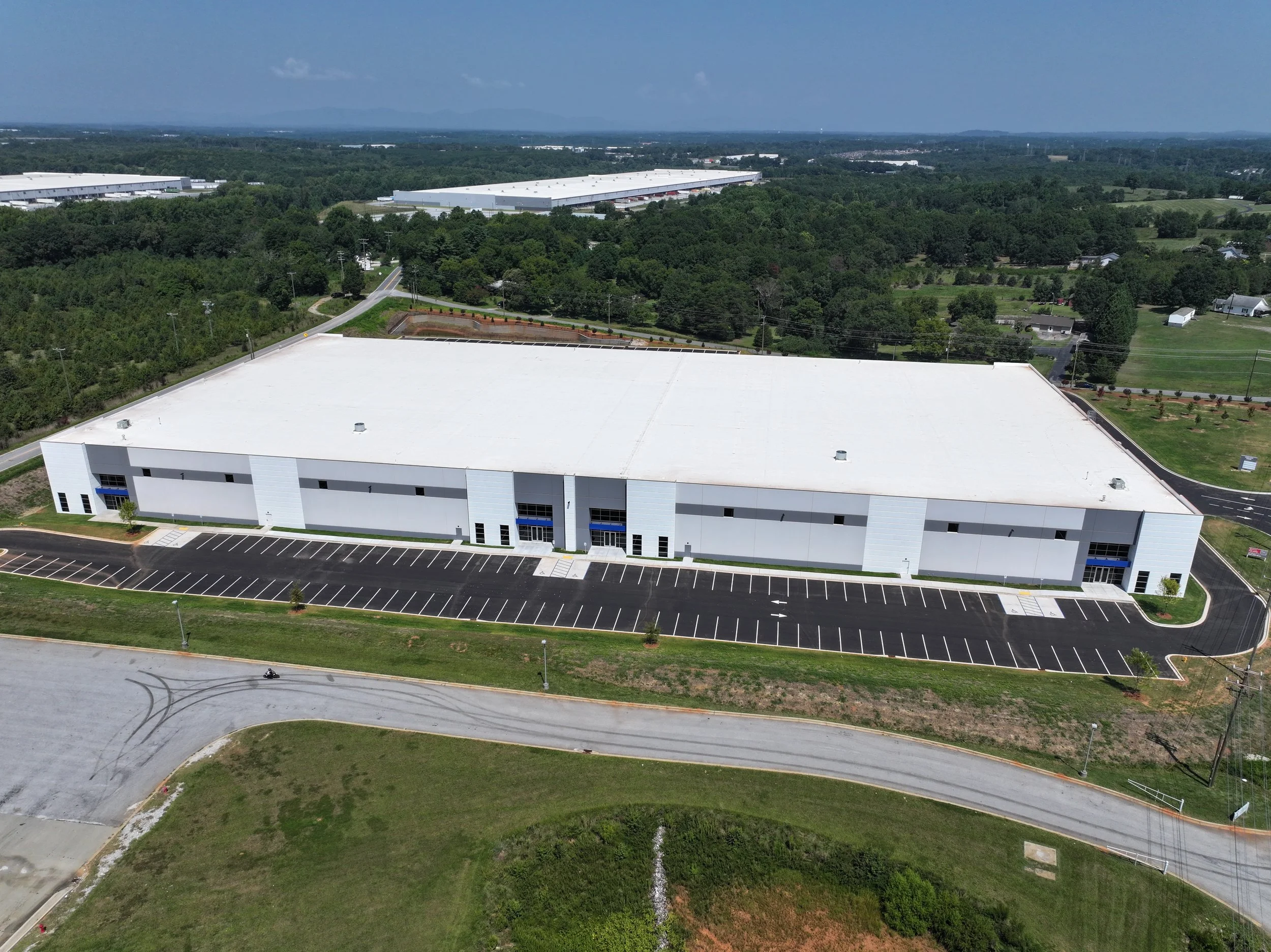 Aerial view of a large industrial warehouse with a white roof, surrounded by parking lots and greenery on a clear day.