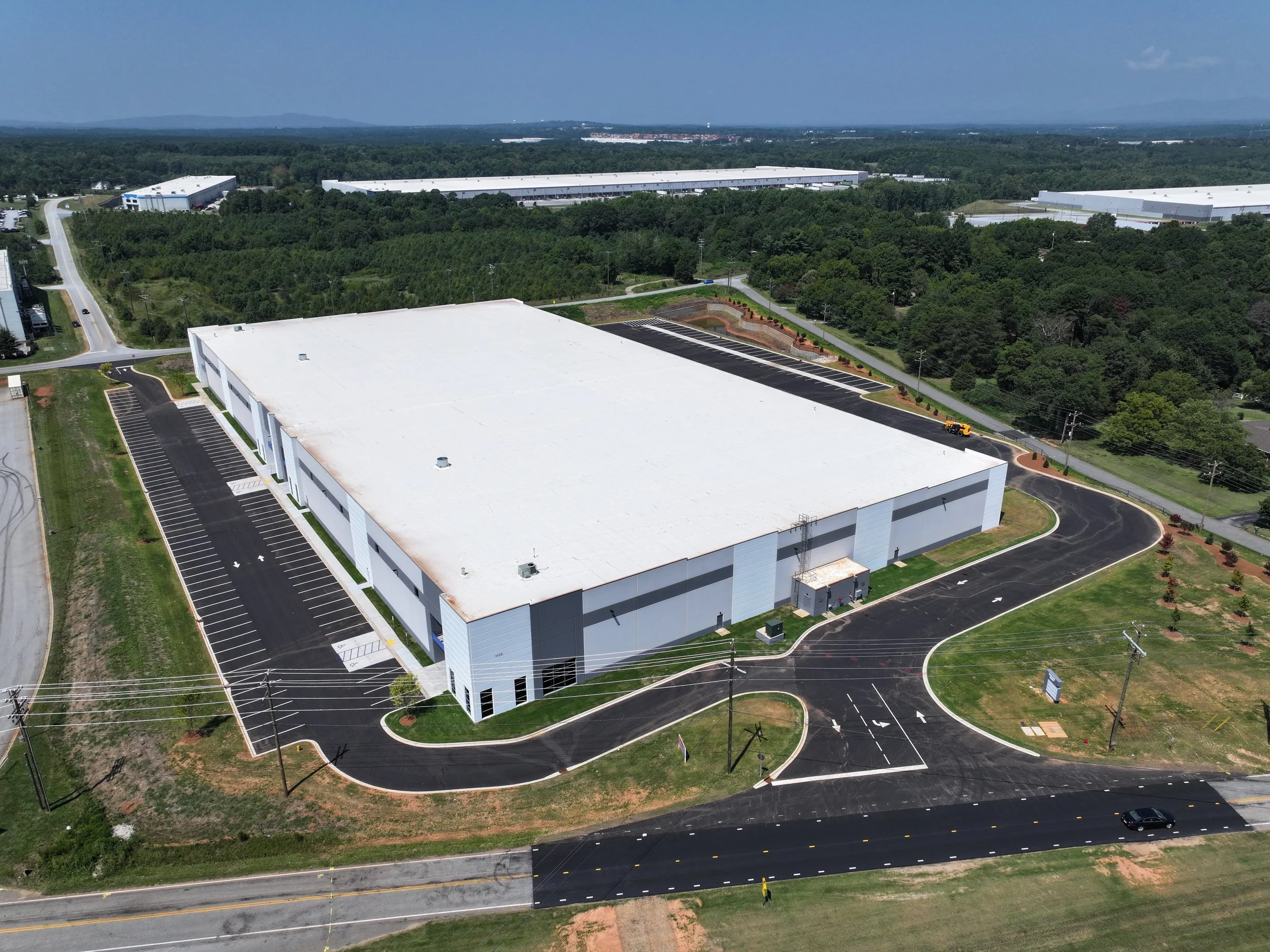 Aerial view of a large industrial warehouse building with a white roof, surrounded by parking lots and roads, located in a green, forested area on a clear day.