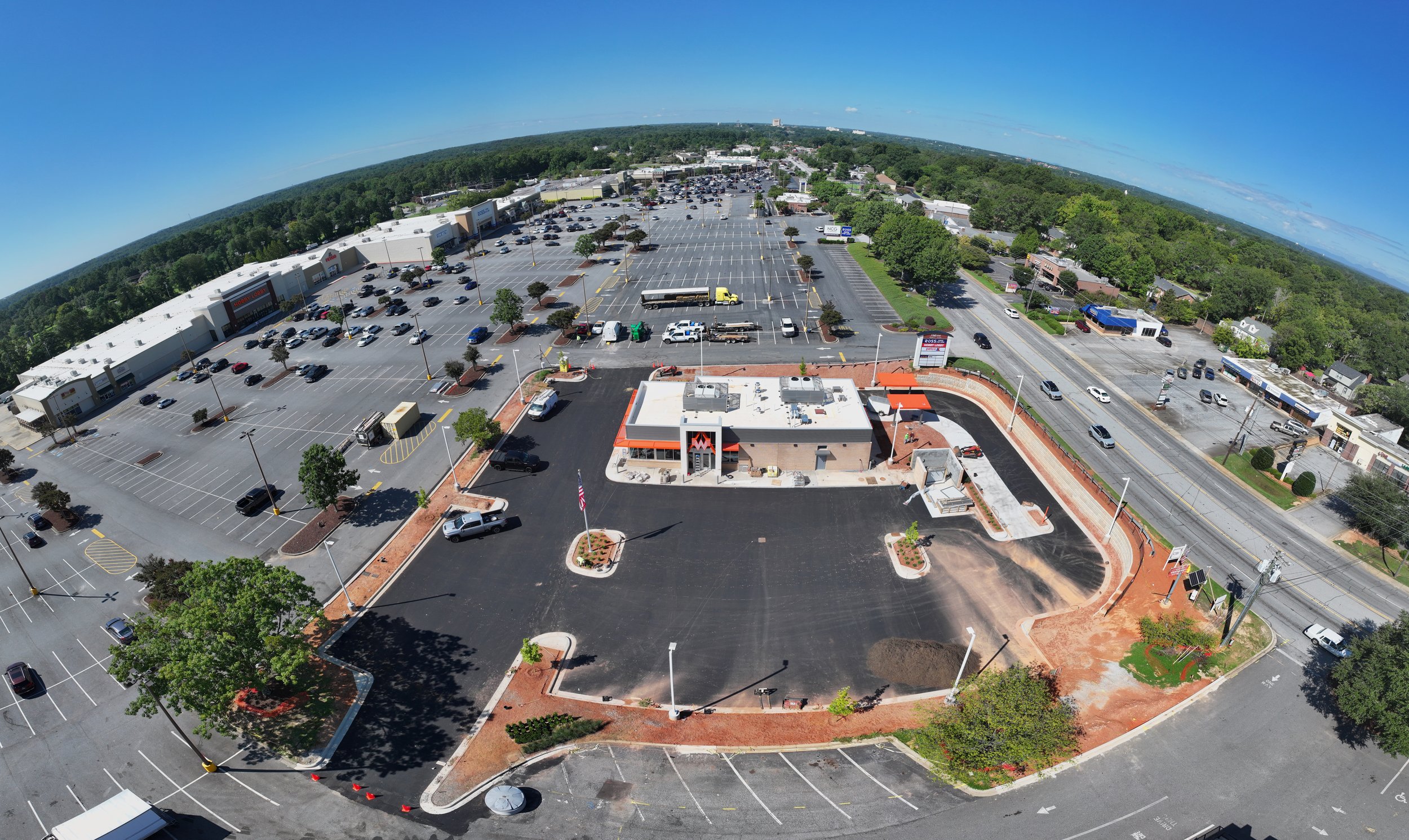 Aerial view of a shopping center with a fast food restaurant. Large parking lot with numerous cars. Surrounding buildings, trees, and a main road with vehicles in the distance. Clear blue sky.