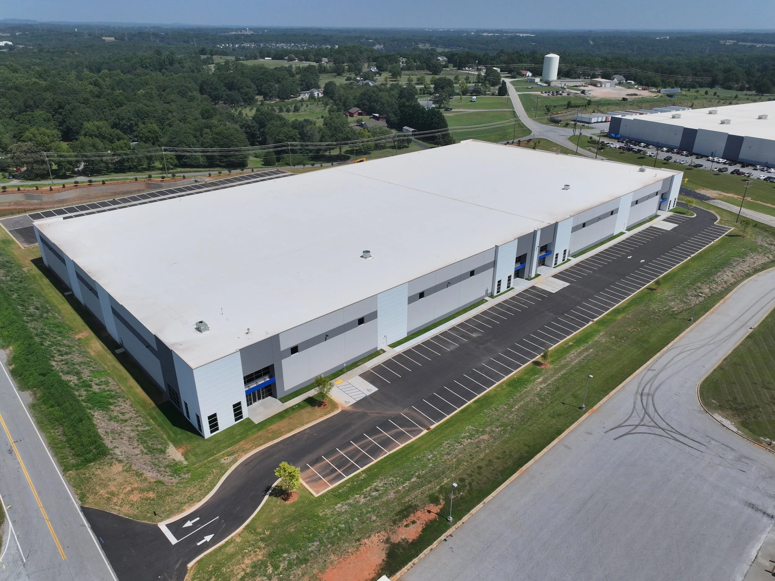 A large, white-roofed warehouse with a parking lot, surrounded by greenery and roads.