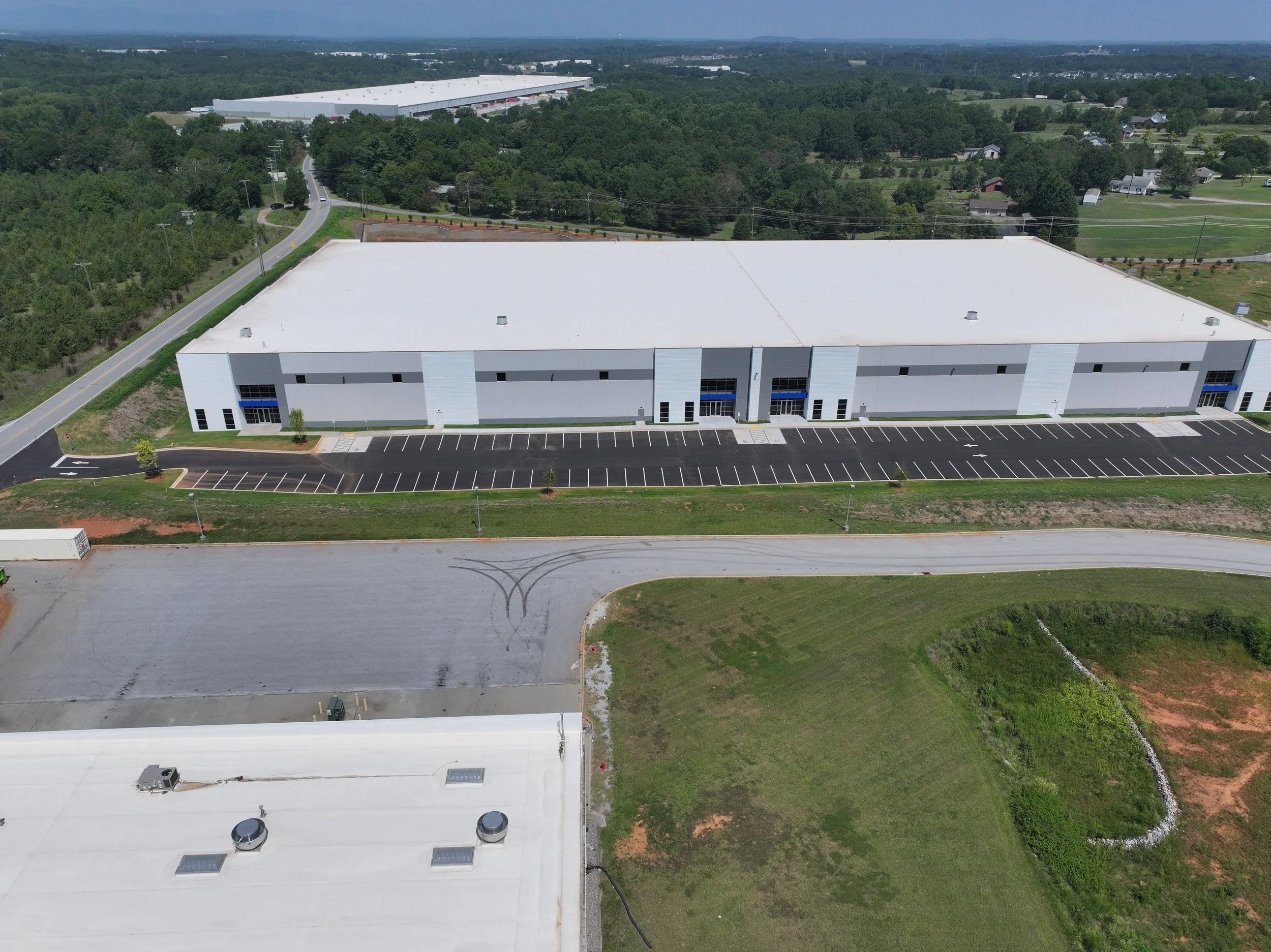 Aerial view of a large white industrial building with an adjacent parking lot, surrounded by green grass and trees, under a clear sky.