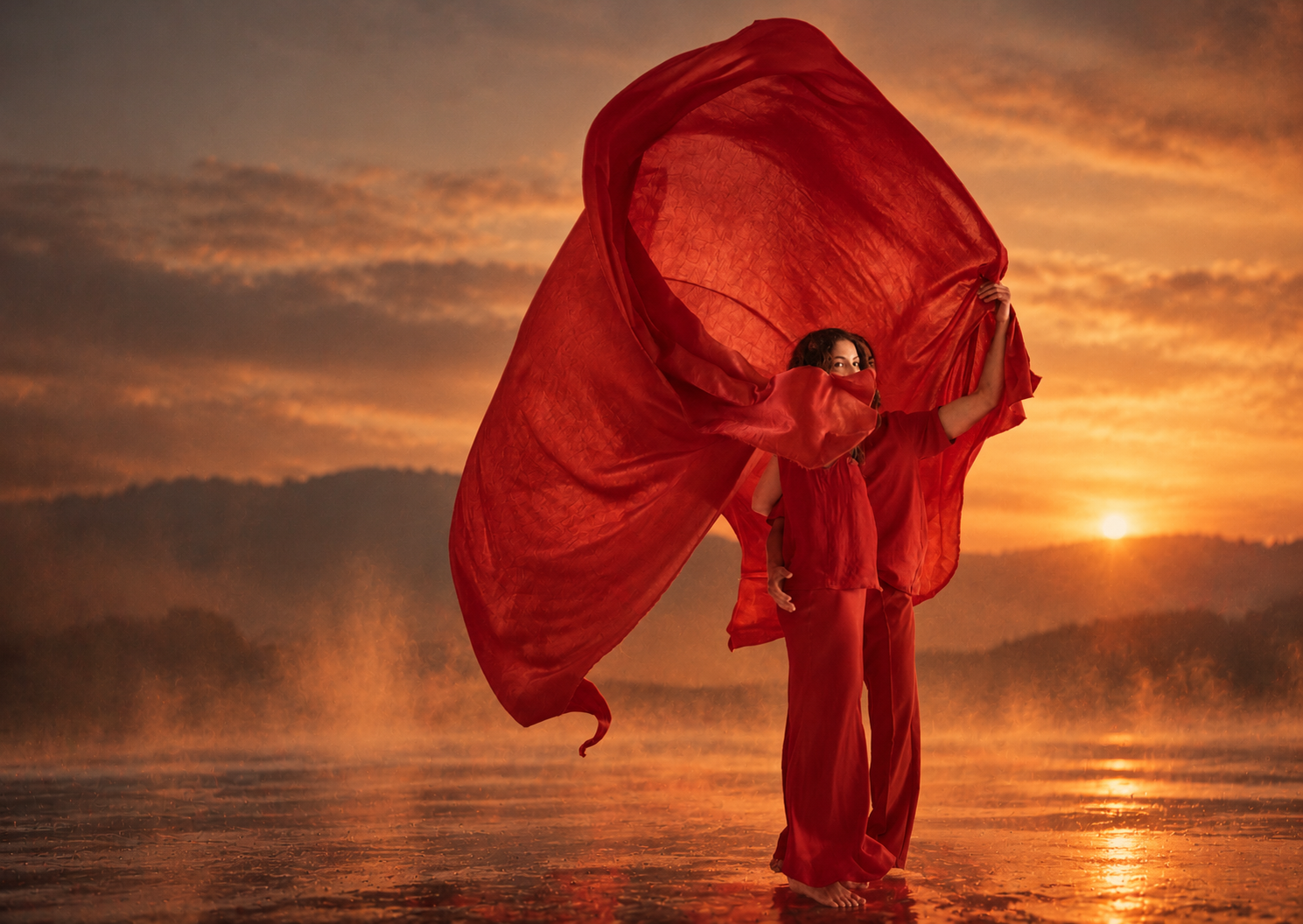 Woman in red clothing holding billowing fabric on a beach at sunset