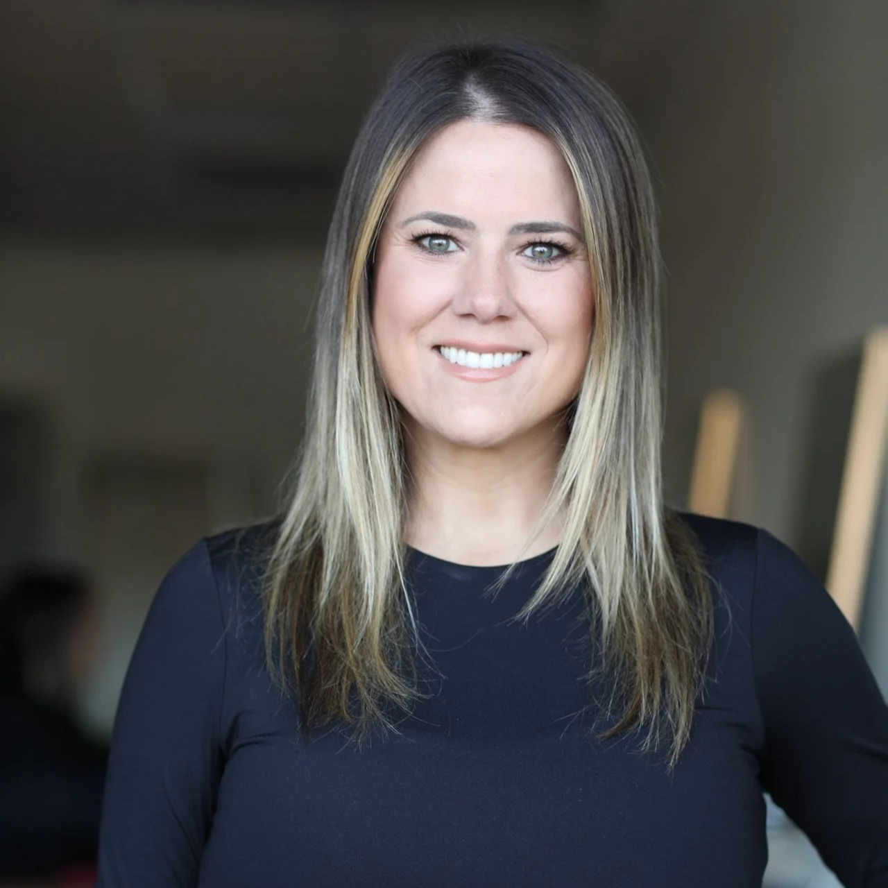 A woman with long, straight blonde hair and blue eyes smiling at the camera, wearing a black top, in a blurred indoor setting.