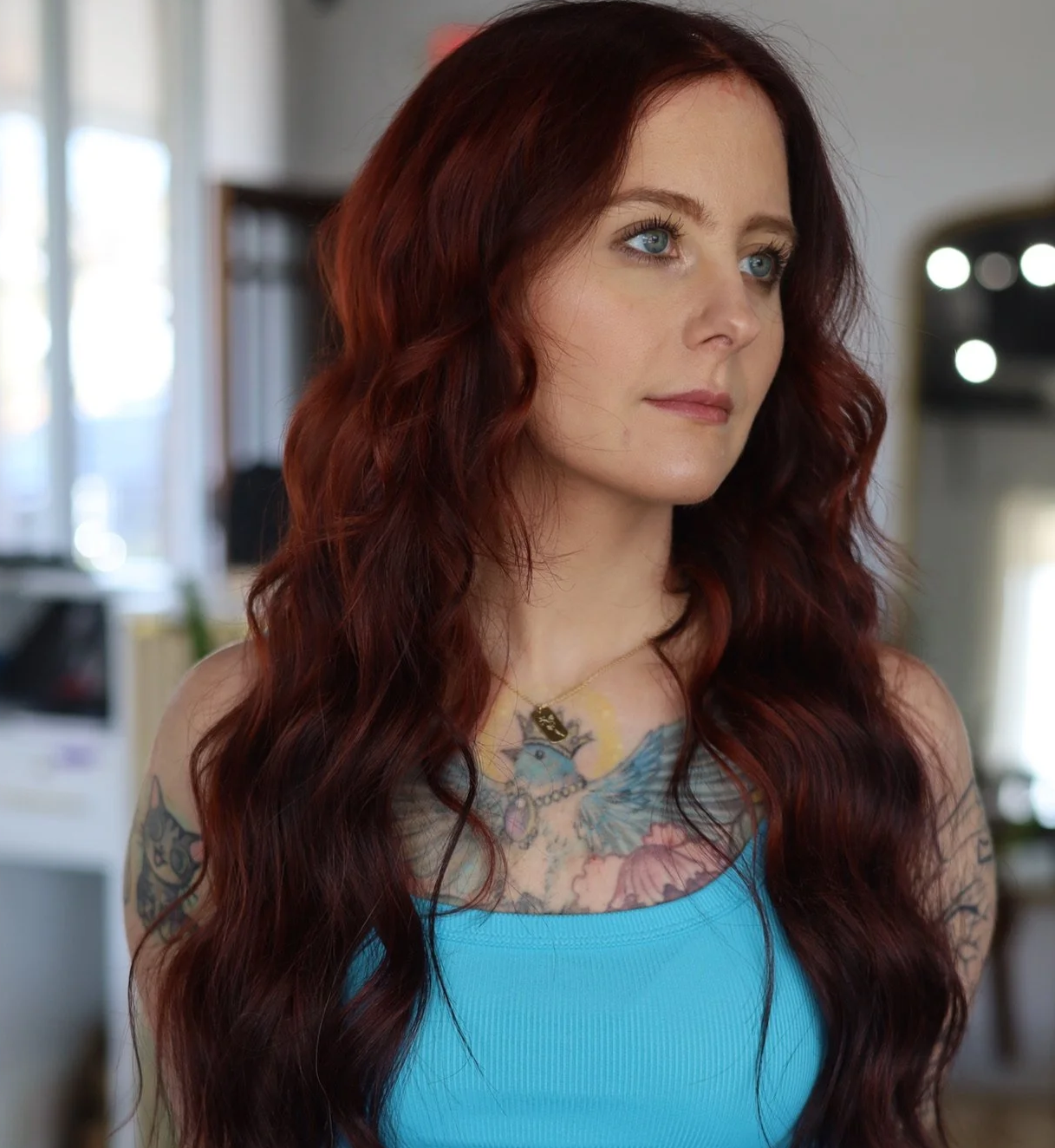 A woman with long, wavy red hair and blue eyes, wearing a blue top and a necklace with a dog tag, standing indoors with tattoos on her chest and arms, and a blurred background of a kitchen and windows.