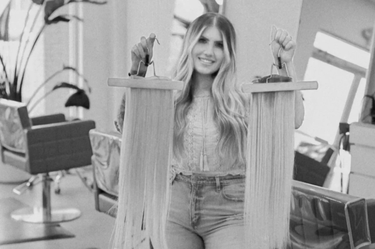 A woman with long, wavy blonde hair smiling and holding a wooden hanger with photos, standing in a salon or studio with chairs and equipment in the background.