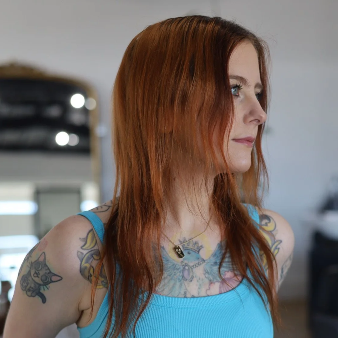 Side profile of a woman with long red hair, tattoos on her shoulders and chest, wearing a blue tank top, indoors.