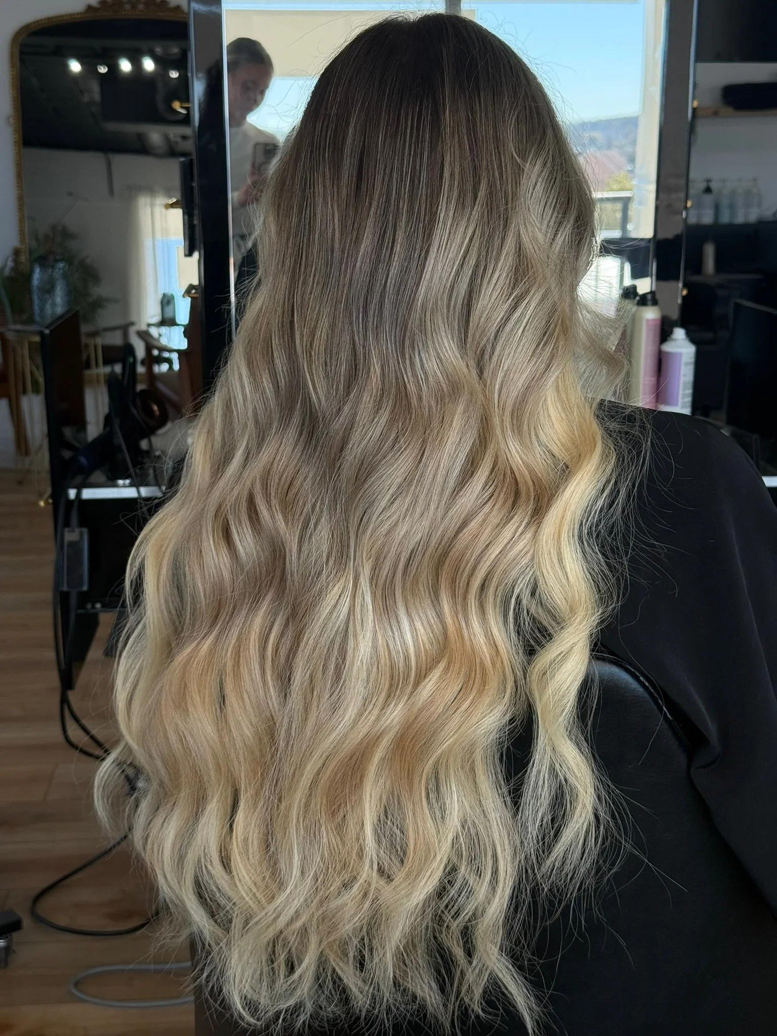 Long wavy blonde hair in a salon setting, with a mirror and various hair products visible in the background.