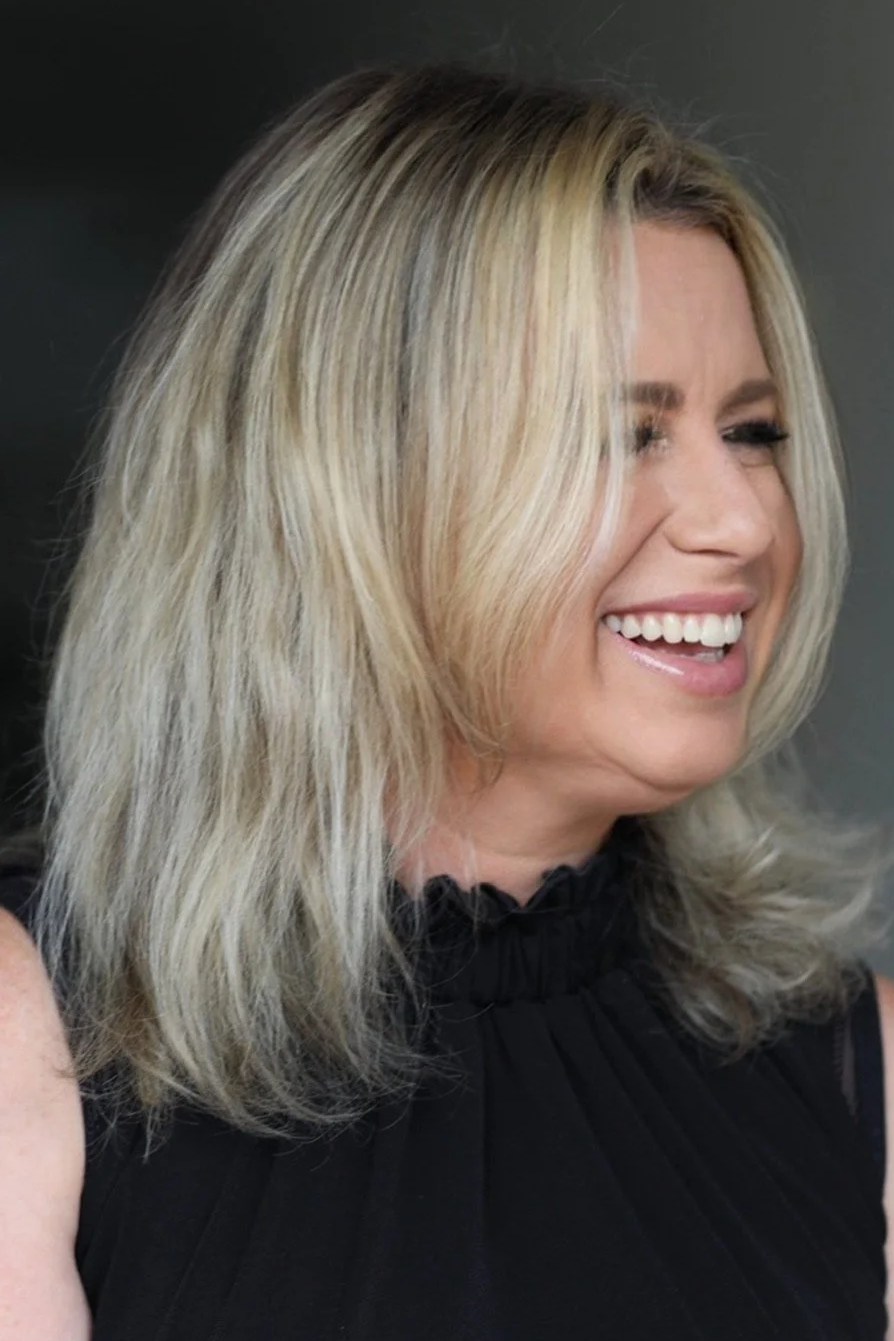 A woman with shoulder-length blonde hair, wearing a black sleeveless top, smiling and looking to the side against a dark background.