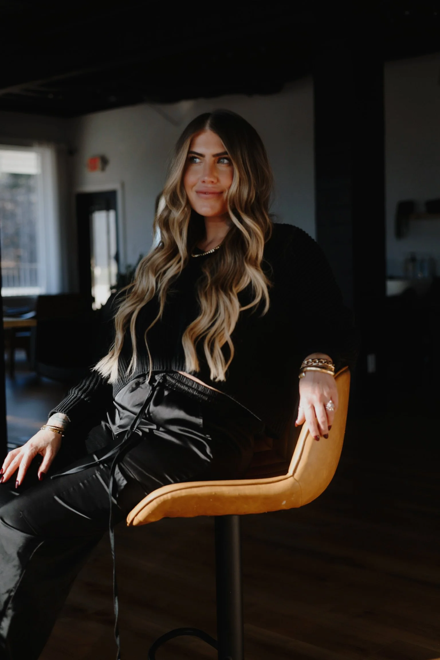 A woman with long wavy blonde hair sitting on a tan chair in a dimly lit room, wearing a black sweater and leather pants, with bracelets on her wrist and a ring, looking to the side.