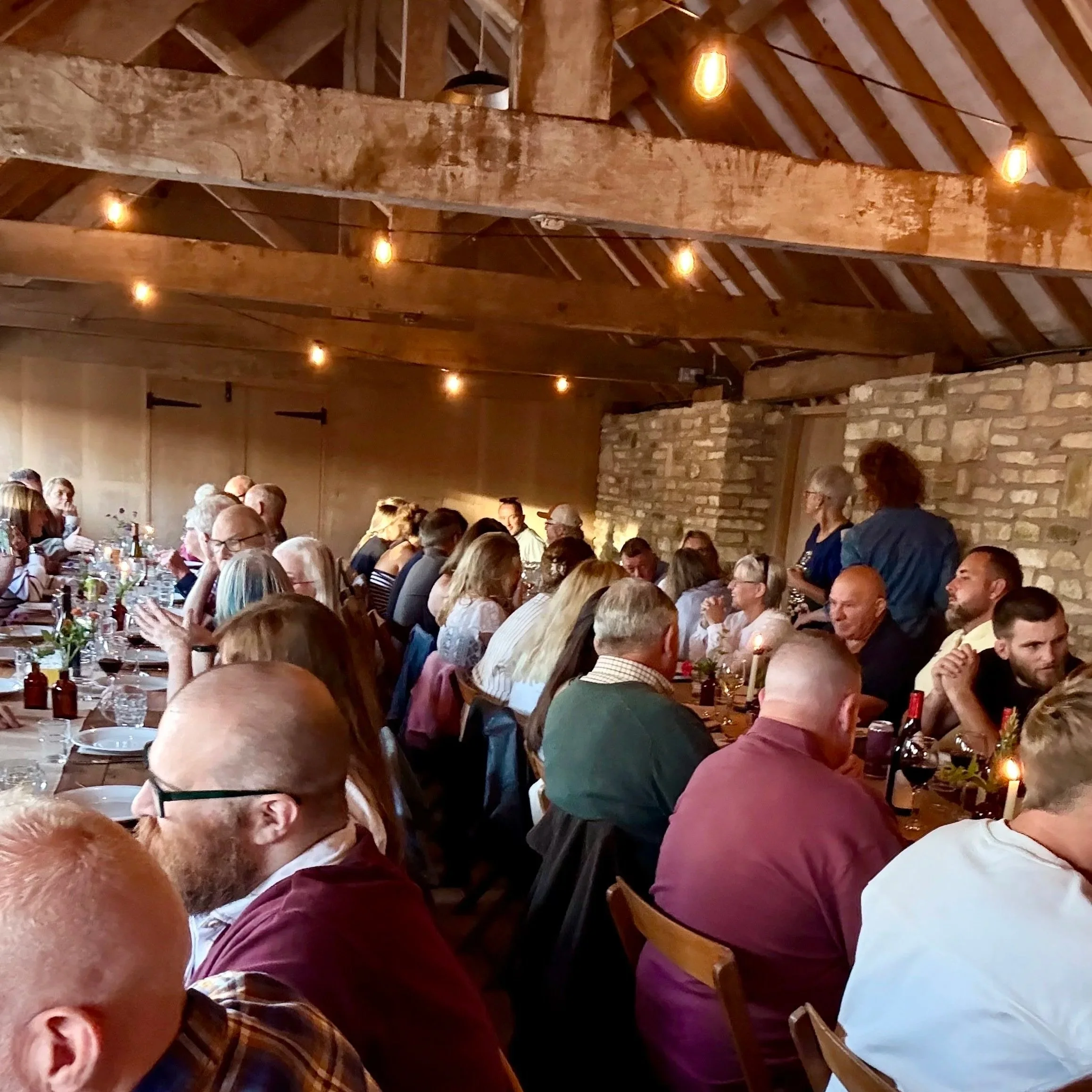 A long communal dining table filled with people in a rustic barn setting, with exposed wooden beams, stone walls and warm hanging lights, as guests eat, drink and chat closely together.