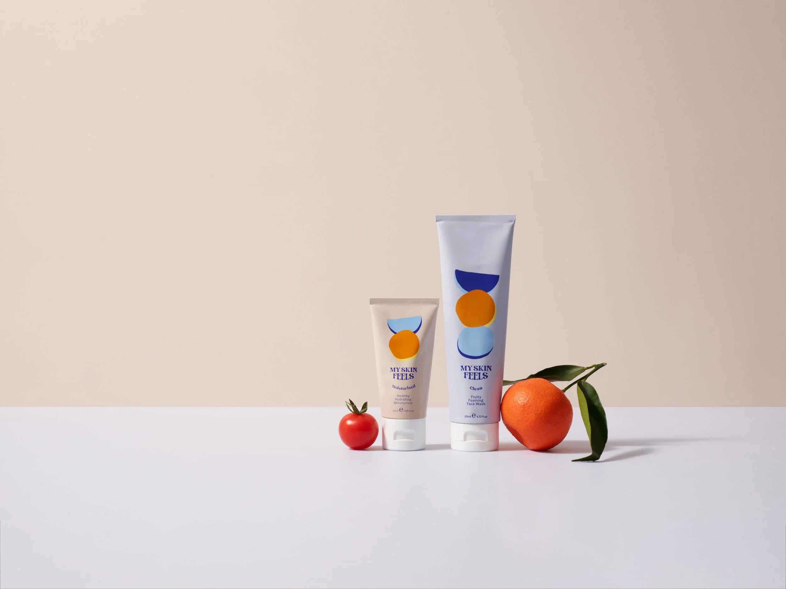 Shot of two My Skin Feels products with a tomato and an orange against a peach background.