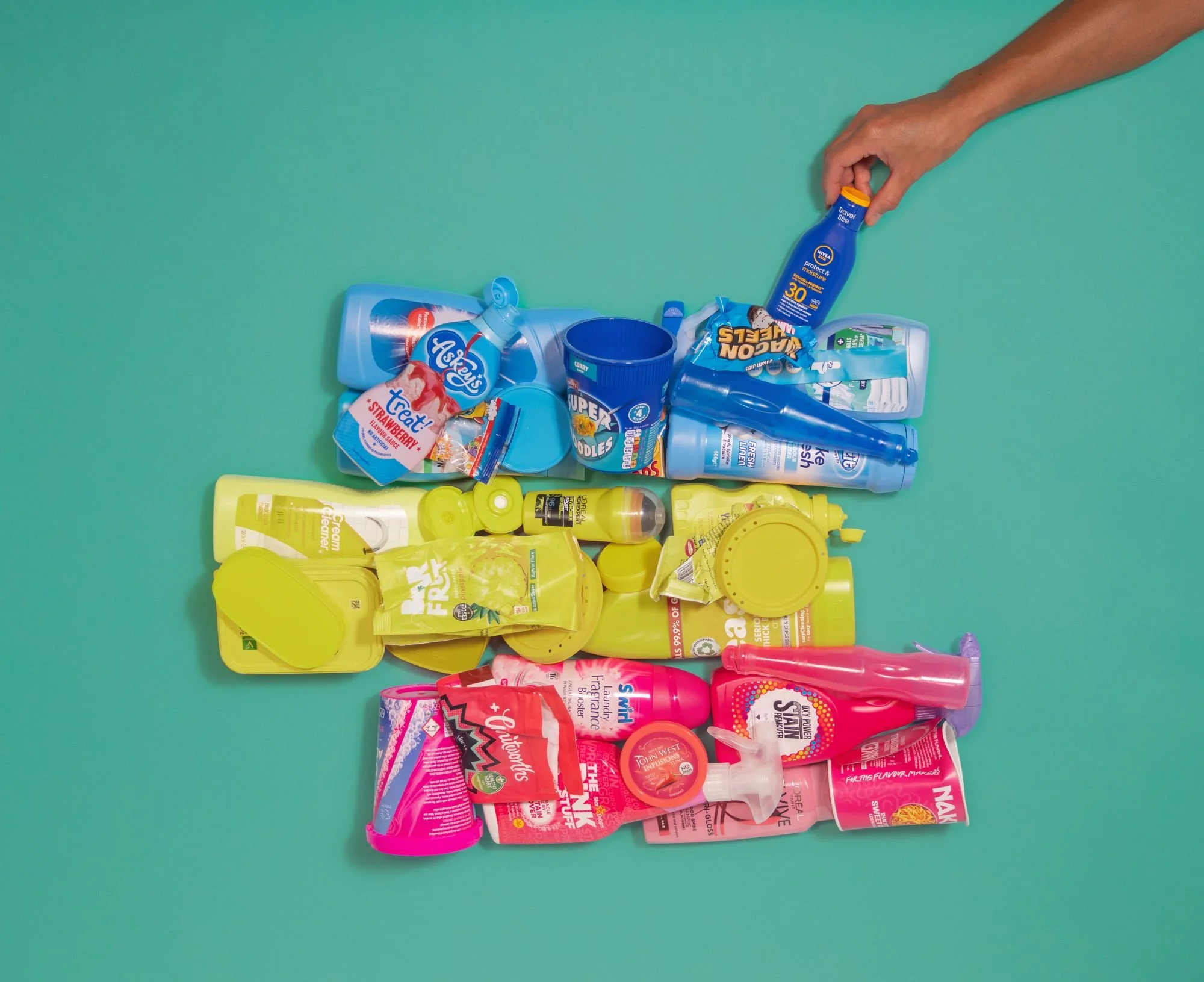 Overhead shot of empty plastic bottles, tubs, wrappers and containers arranged in colour bands of blue, yellow and pink on a green background, with a hand placing a blue bottle at the top.