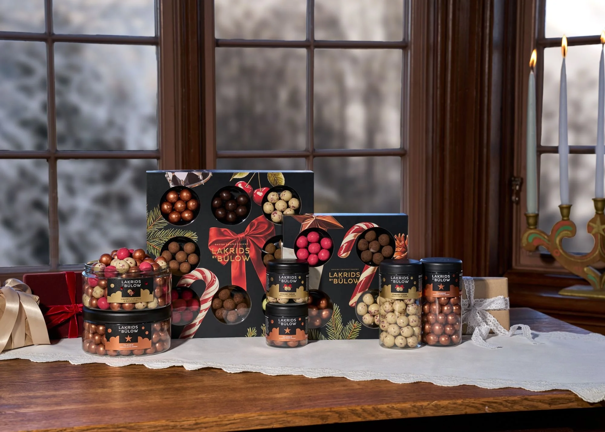 Selection of Lakrids by Bülow liquorice gift boxes and tubs displayed on a wooden table, with wrapped presents and lit candles creating a festive winter setting.