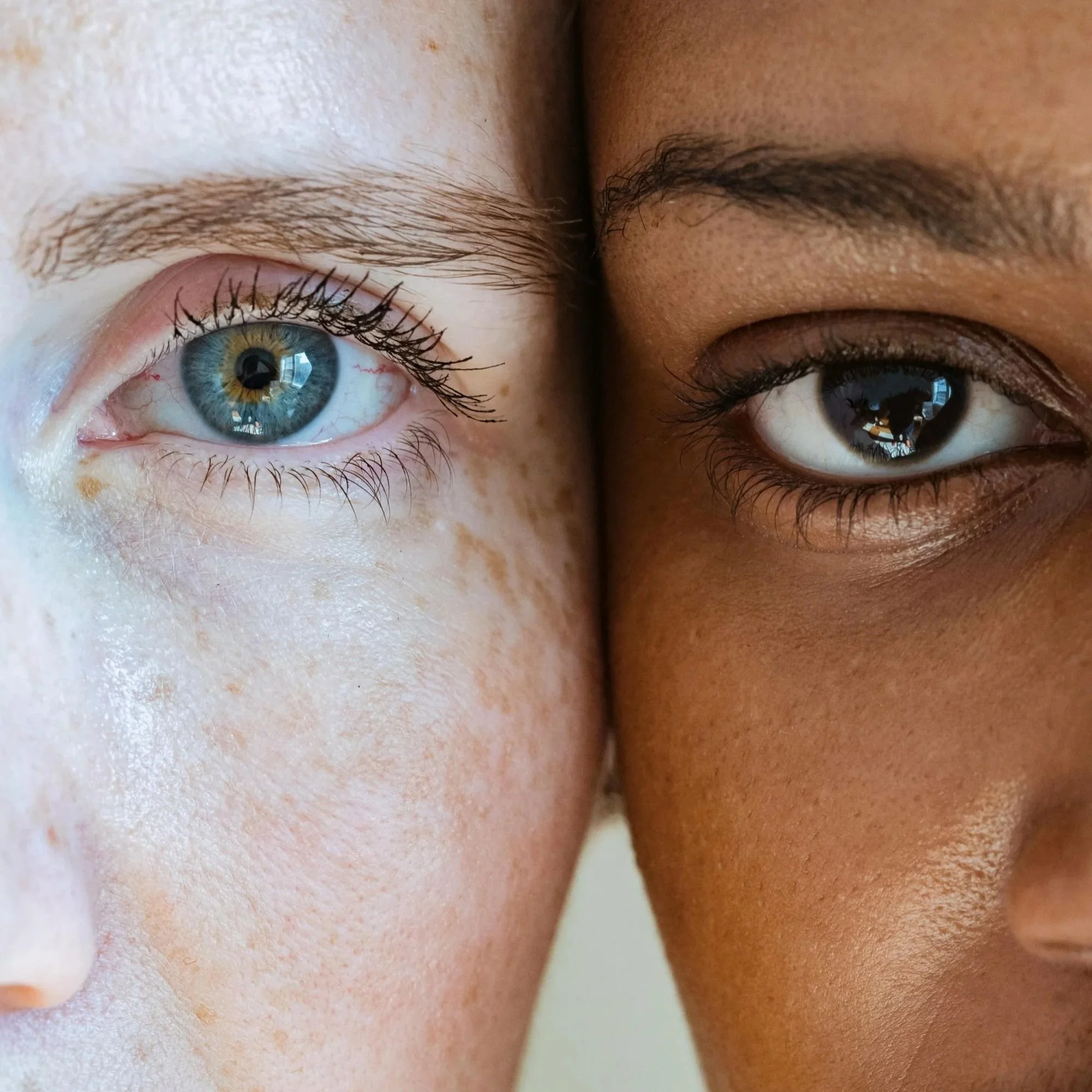 Extreme close-up of two women’s faces side by side, cropped down the middle; one has light skin with freckles and a blue eye; the other has dark brown skin and a brown eye.