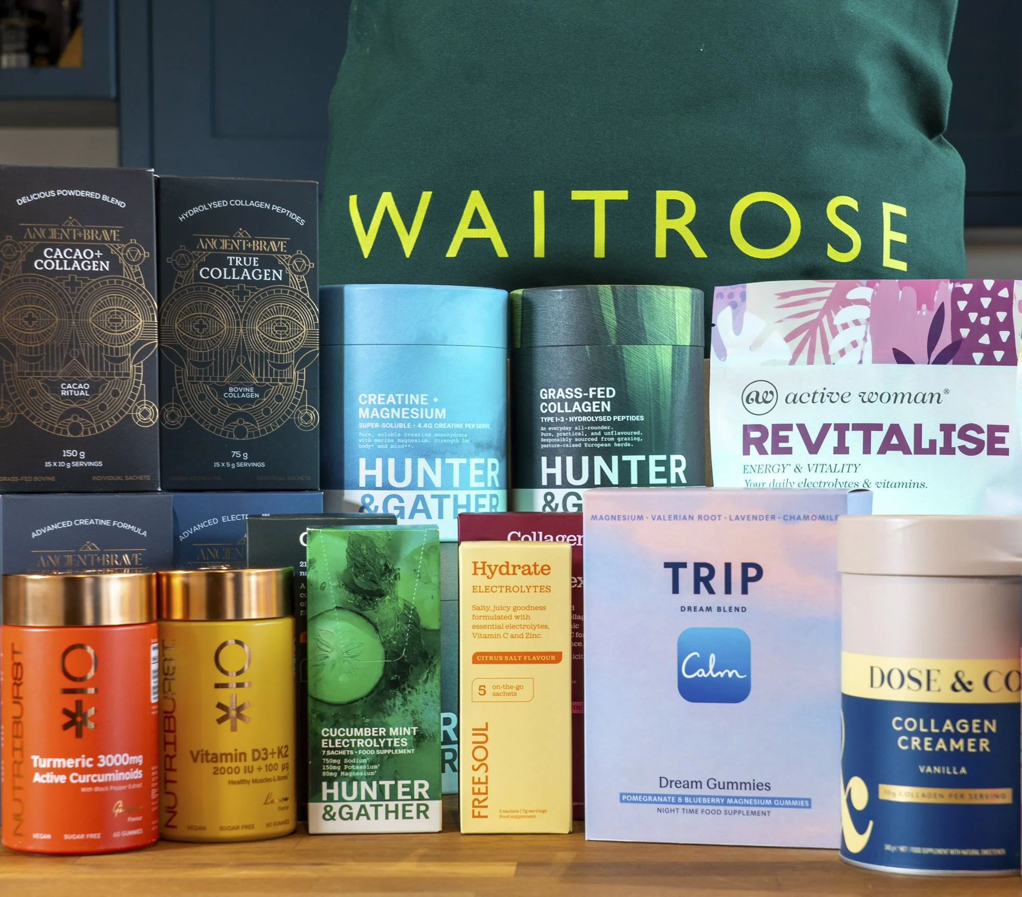 A green Waitrose bag behind products from brands including Ancient + Brave, Hunter & Gather, Active Woman, NutriBurst, TRIP, Dose & Co and Free Soul.