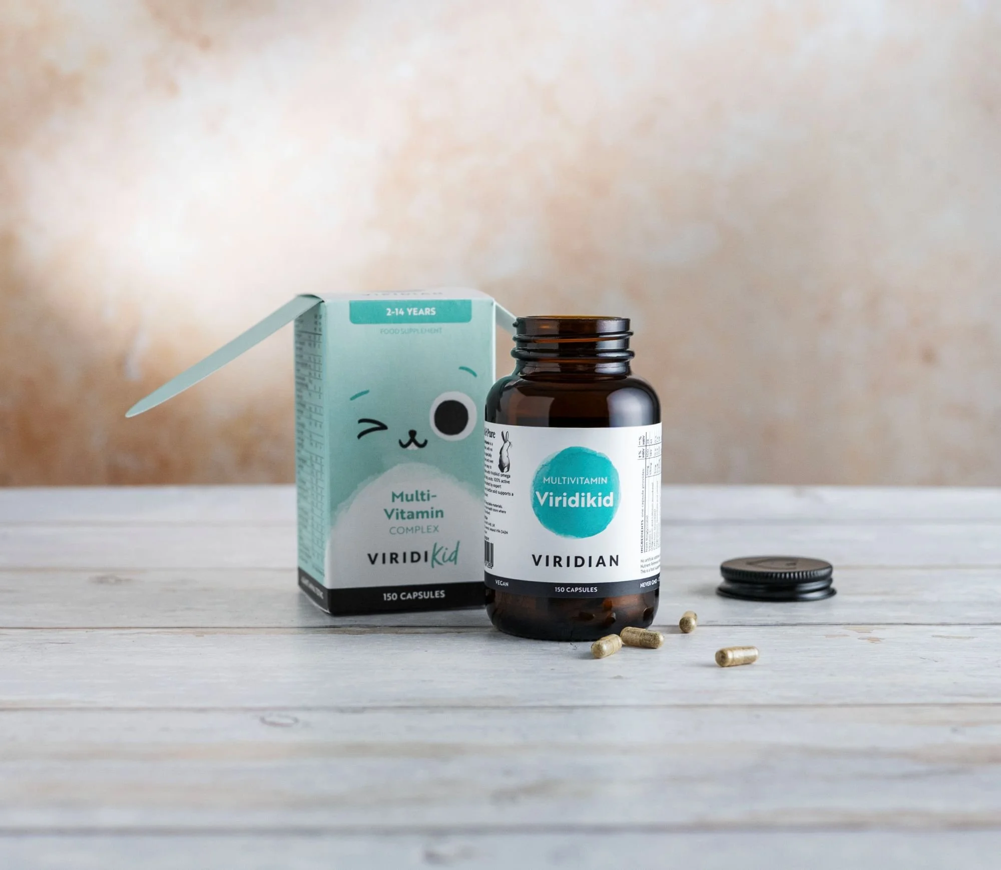 Viridikid Multivitamin bottle next to its light blue box with a few scattered capsules. on a wooden surface.
