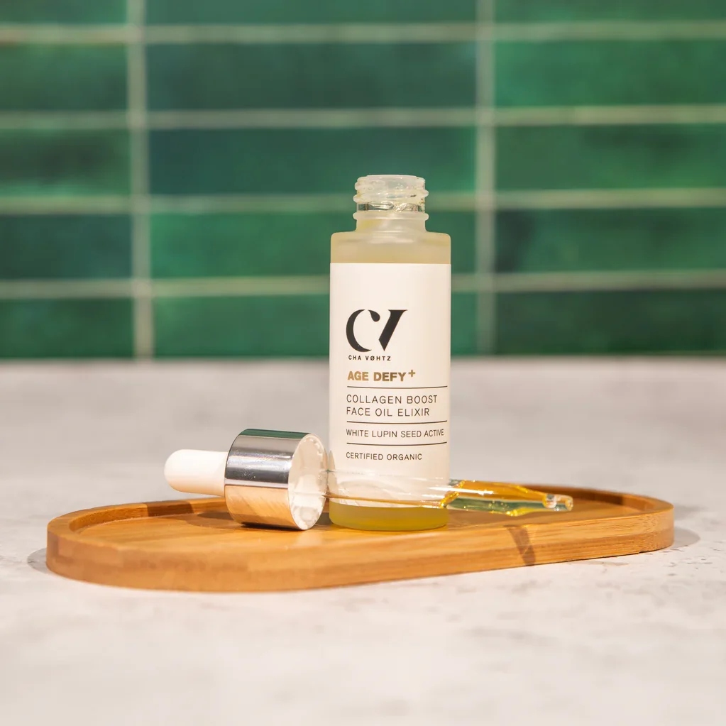 Glass bottle of facial oil with dropper, labelled “Age Defy+ Collagen Boost Face Oil Elixir,” resting on a wooden tray against a green tiled background.