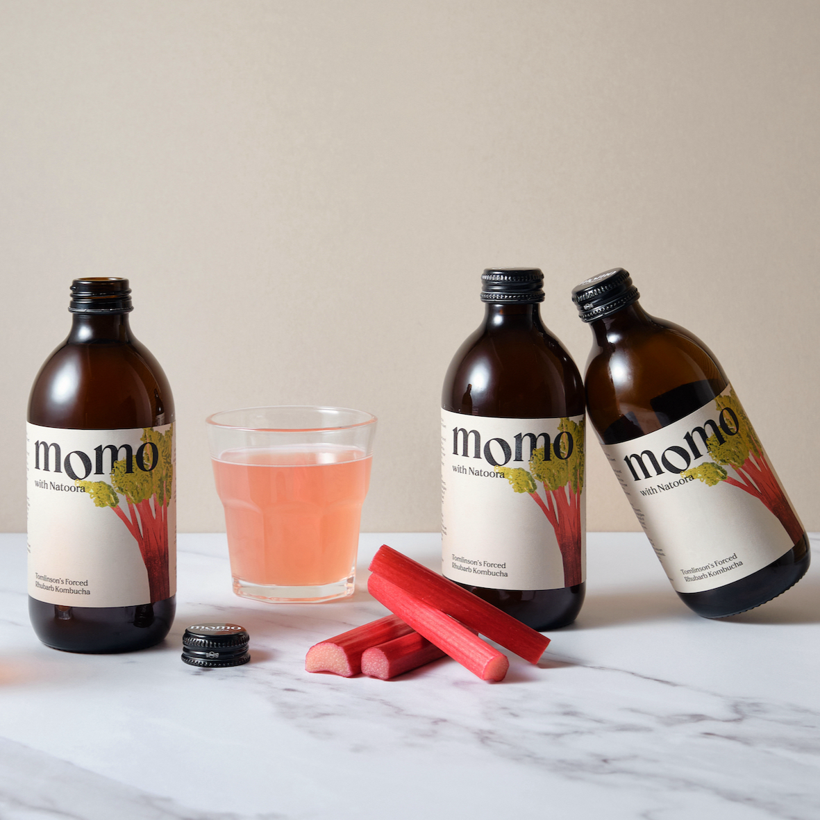 A glass of Momo with Natoora Rhubarb Kombucha along with three bottles of the drink and some sticks of rhubarb on a white marble surface.