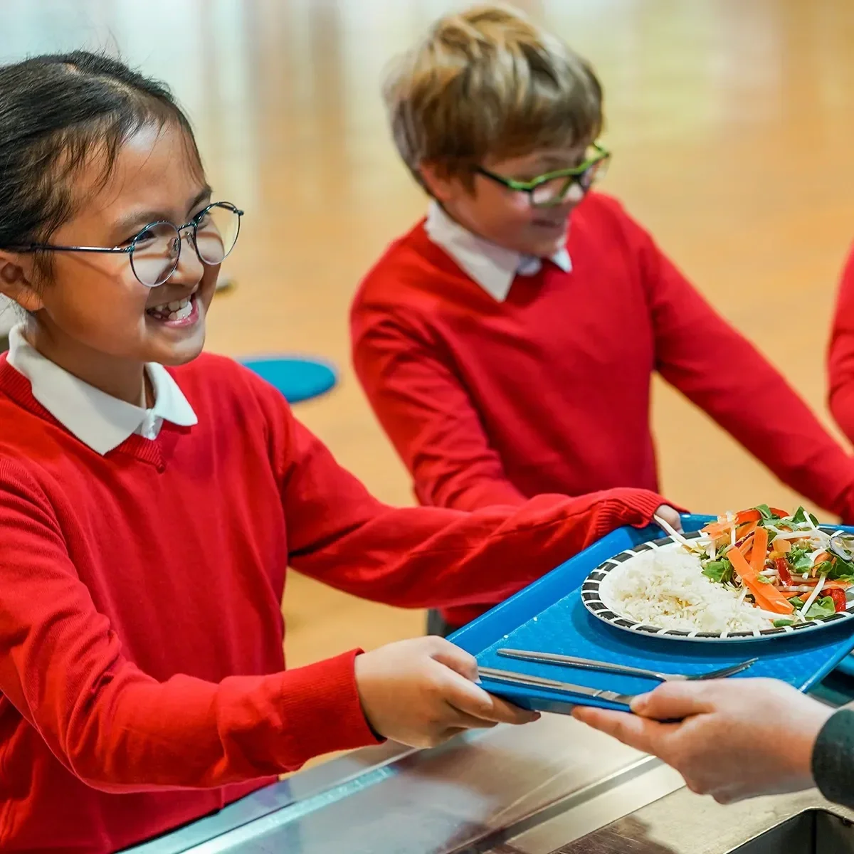 Schoolchildren in red uniforms smiling as they receive trays of food from a server in a cafeteria, with plates of rice and vegetables visible.