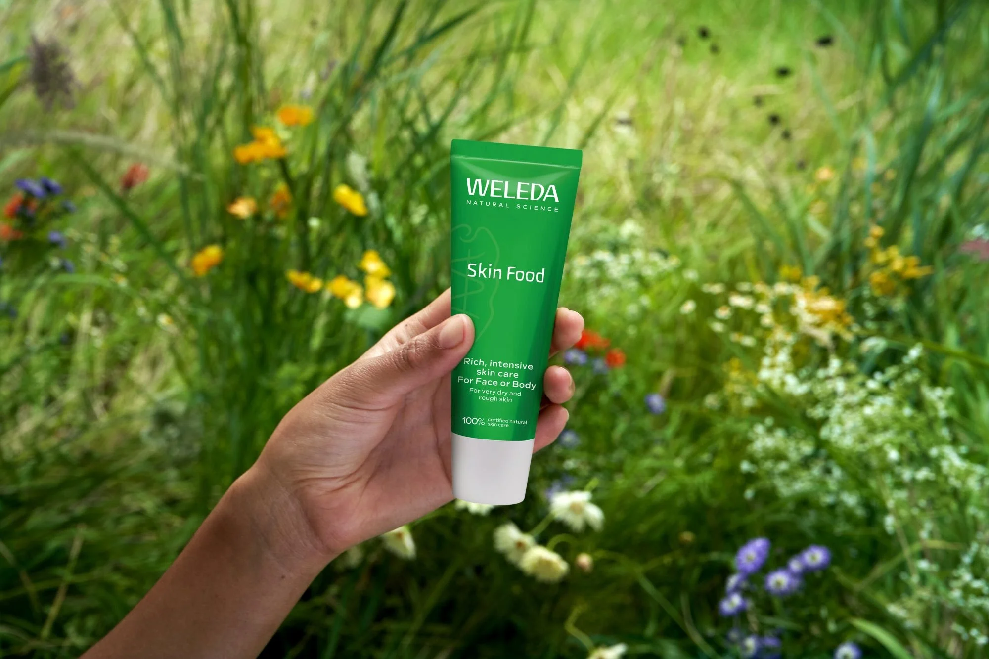 Close up of a hand holding a tube of Weleda Skin Food against a background of wild flowers.