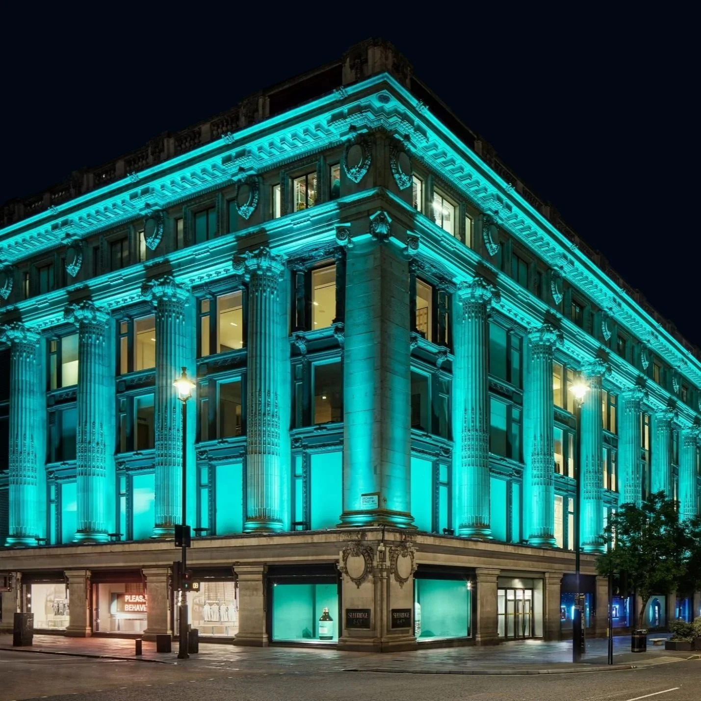 Selfridges department store on Oxford Street at night illuminated in turquoise lighting and lit display windows.