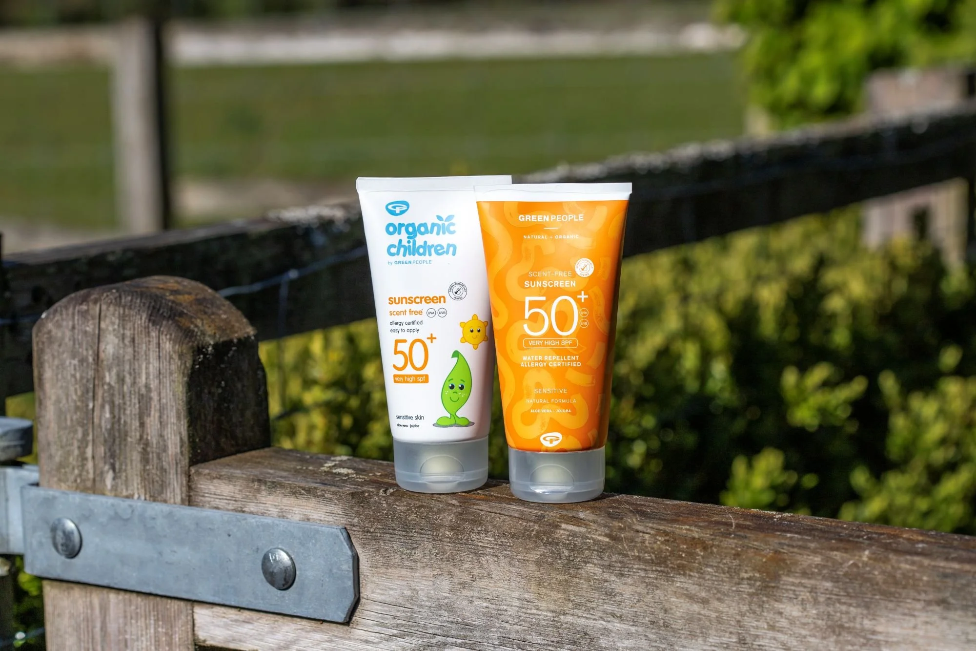 Two Green People SPF50+ sunscreen tubes—one for children and one scent-free—standing on a wooden fence in a sunny outdoor setting.