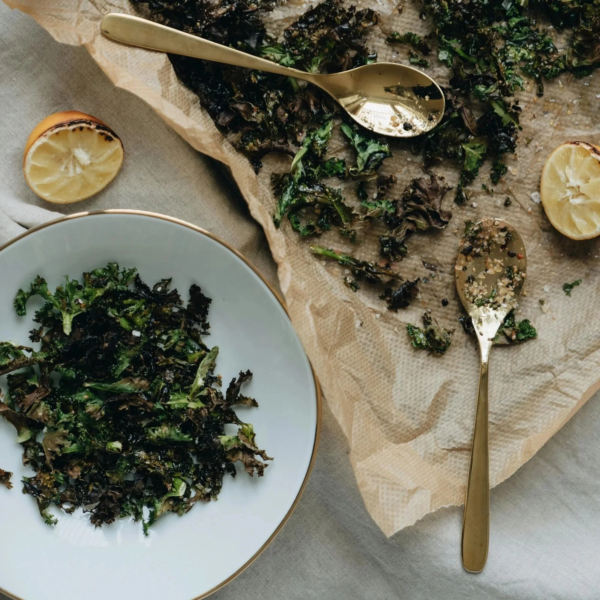 Crispy kale chips on parchment paper with lemon halves and spoons, one serving plated.
