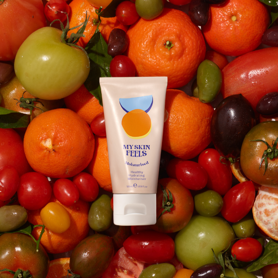 A tube of My Skin Feels moisturiser placed among a vibrant mix of fresh produce, including whole and sliced oranges, cherry and green tomatoes and grapes.