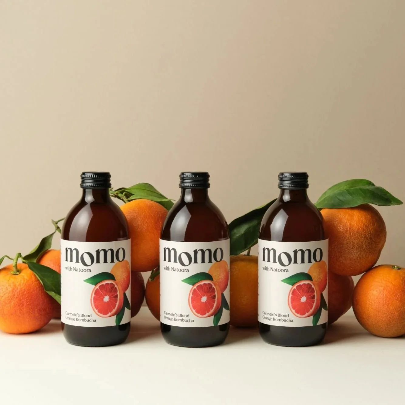 Three brown glass bottles of Momo blood orange kombucha arranged in a row, surrounded by whole oranges with green leaves against a neutral background.