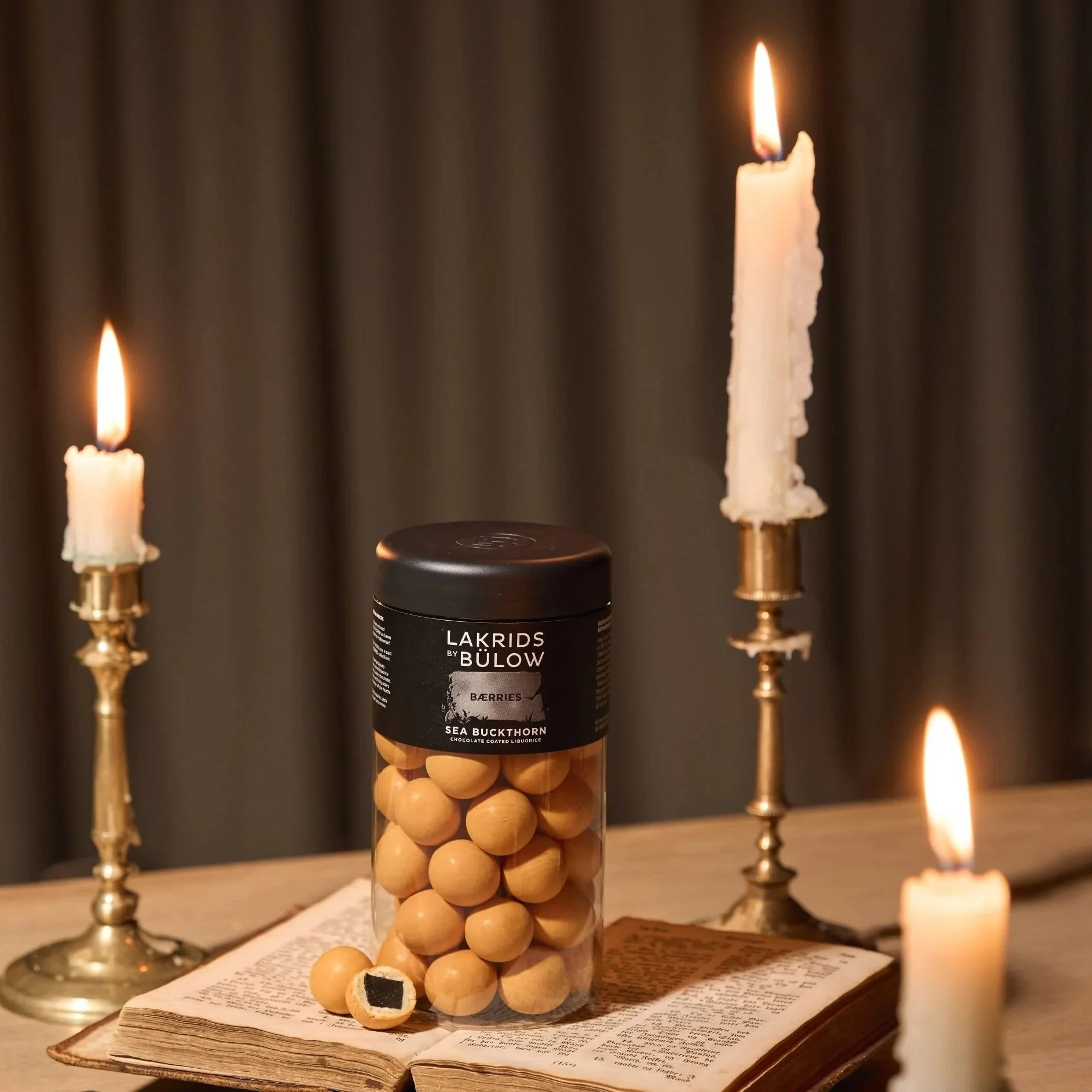 Tub of Lakrids by Bulow Sea Buckthorn  liquorice balls set on an open book with candles around it.