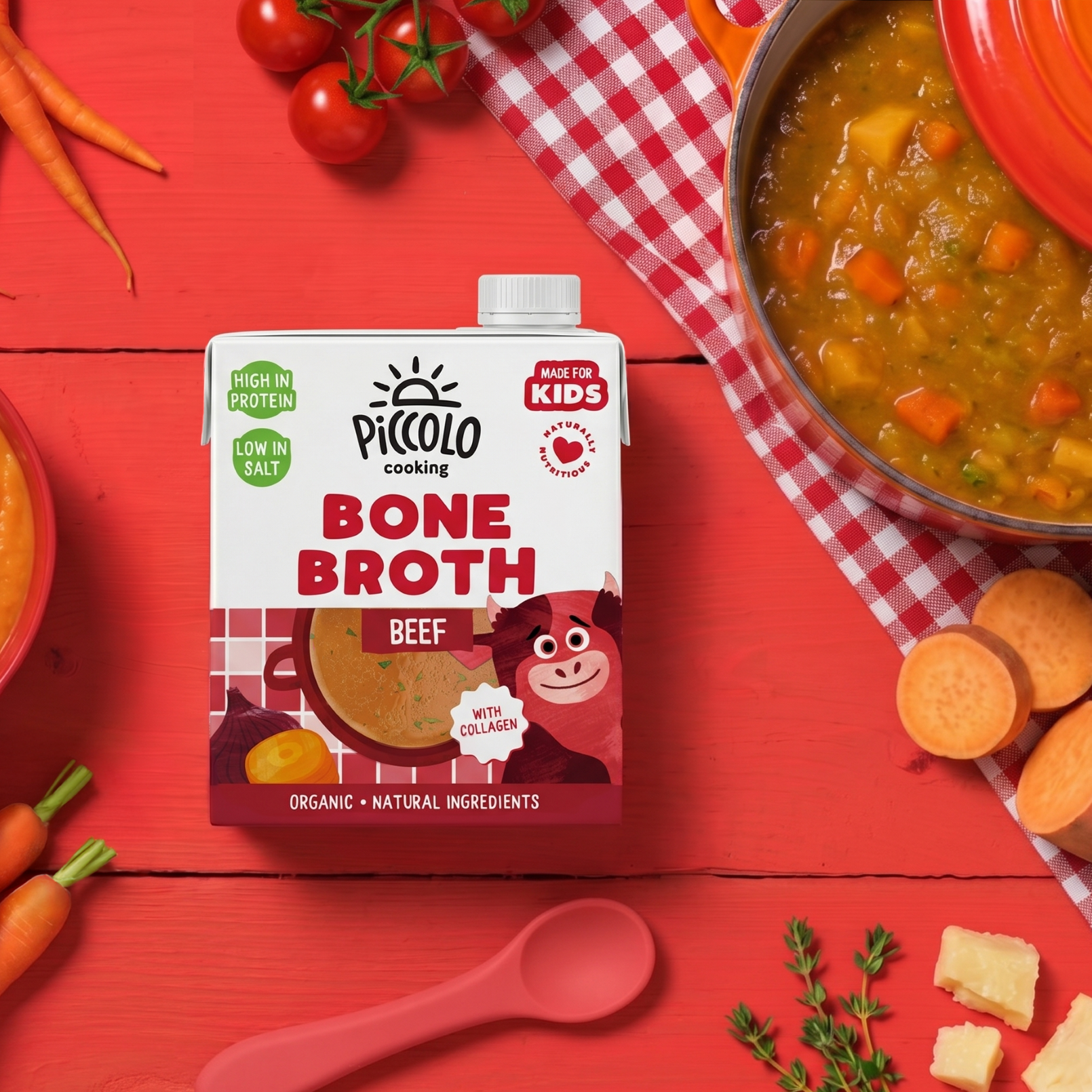 Pack of Piccolo Organic Beef Bone Broth for kids on a red surface, surrounded by carrots, onions, tomatoes and a pan of hearty soup.