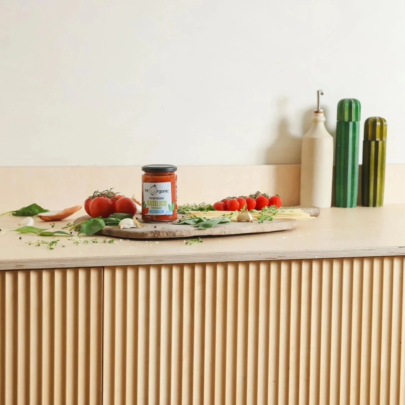 Jar of Mr Organic Basilico pasta sauce on a wooden kitchen counter, surrounded by fresh tomatoes, garlic, herbs and uncooked pasta, with oil bottles in the background.