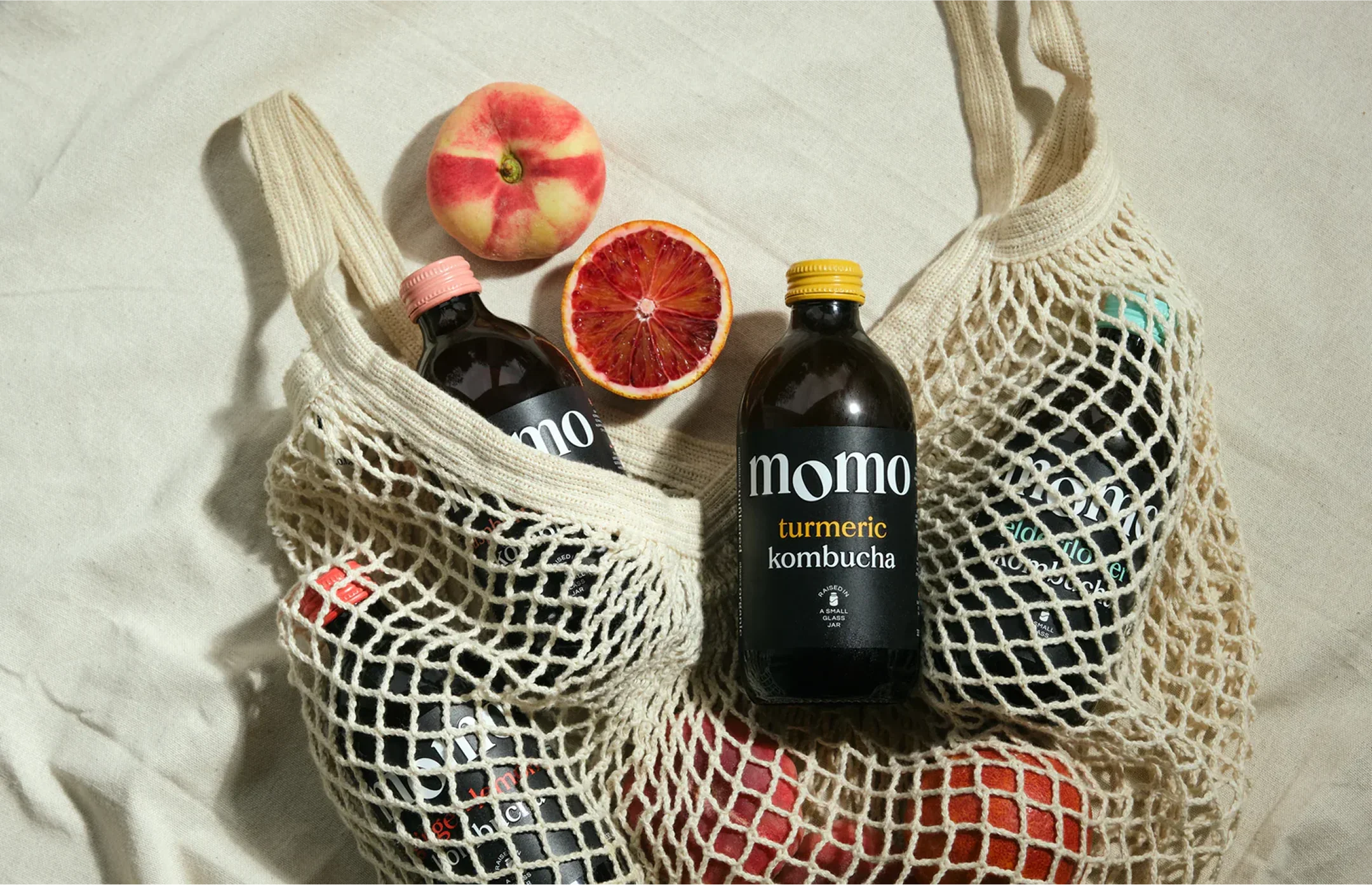 Bottles of Momo kombucha in a string bag with peaches and oranges