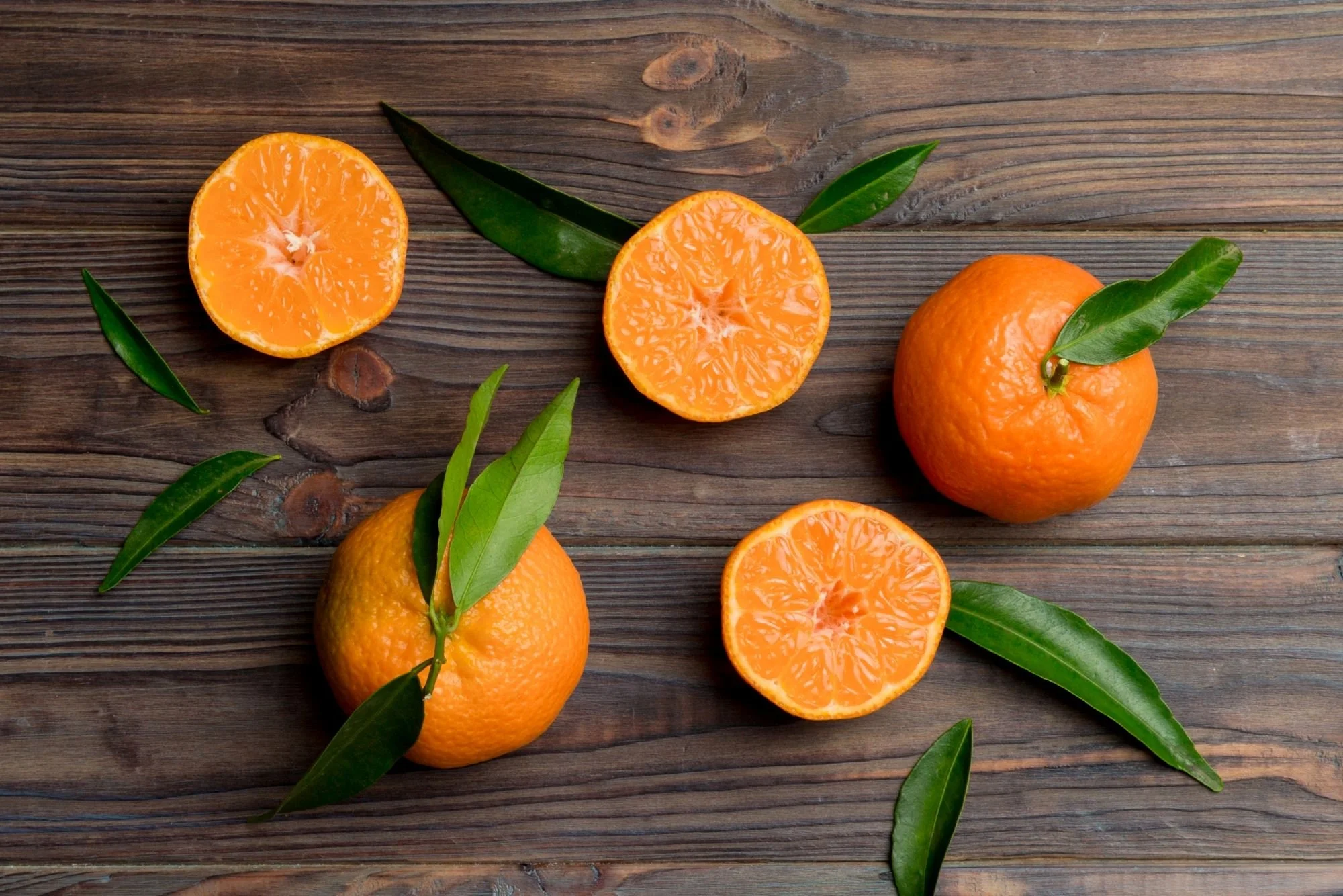 Whole and halved oranges with green leaves arranged on a rustic wooden surface, showing the bright, juicy flesh and textured peel.