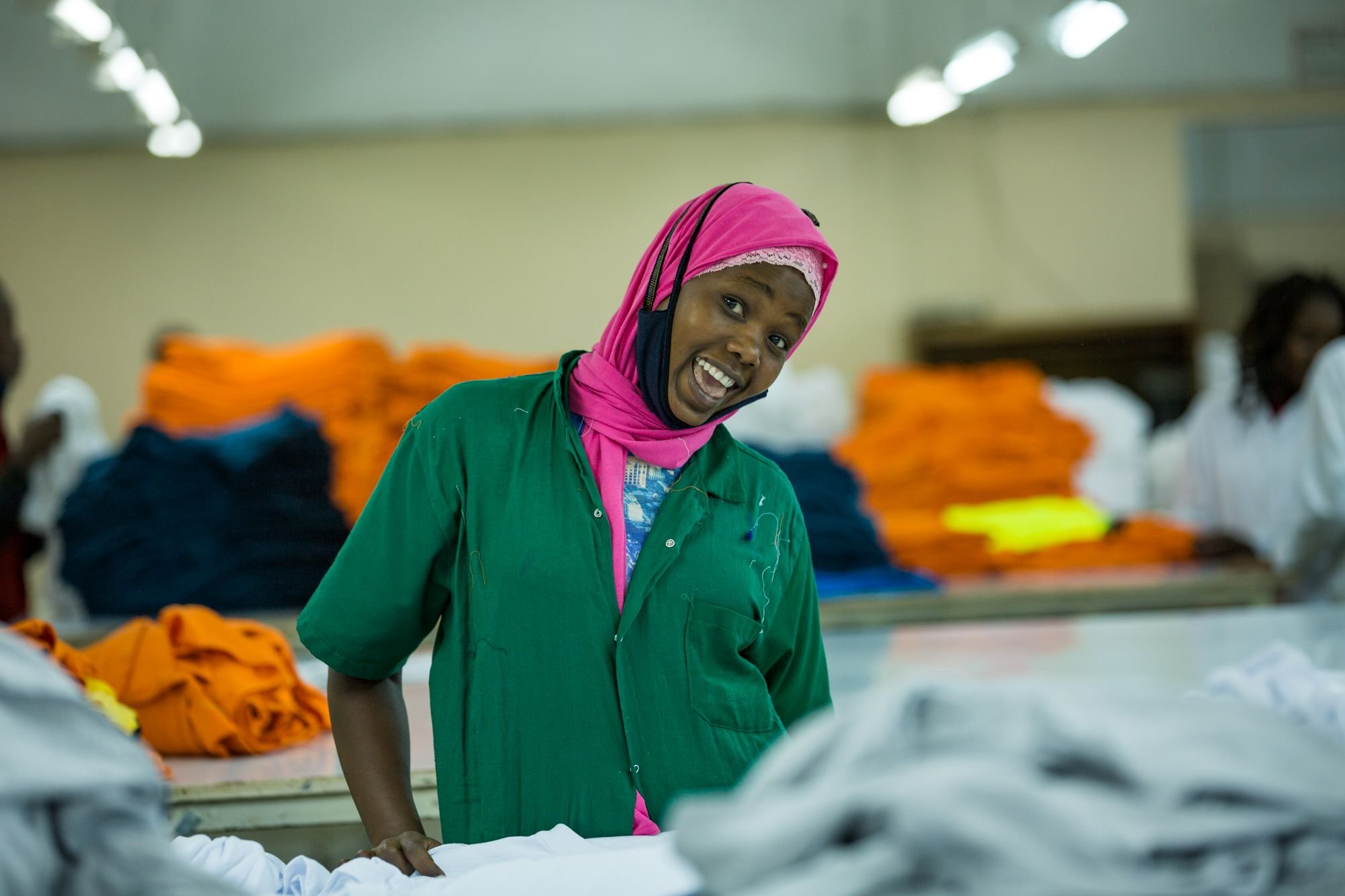 A smiling textile worker in a green uniform and pink headscarf stands at a table in a factory, with stacks of brightly coloured folded garments in the background.